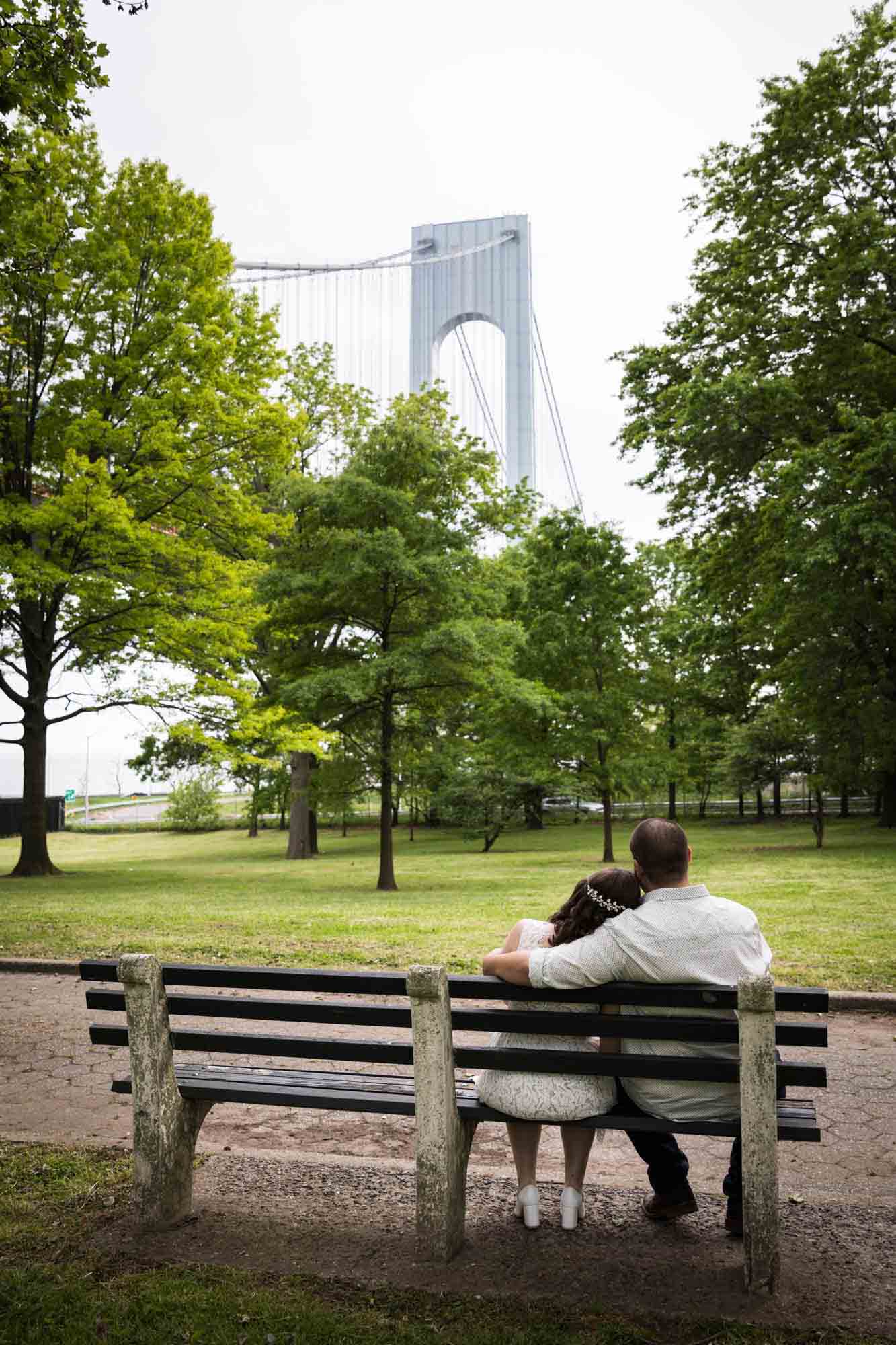 Couple hugging on bench with Verrazano Bridge in background during a Bay Ridge engagement shoot