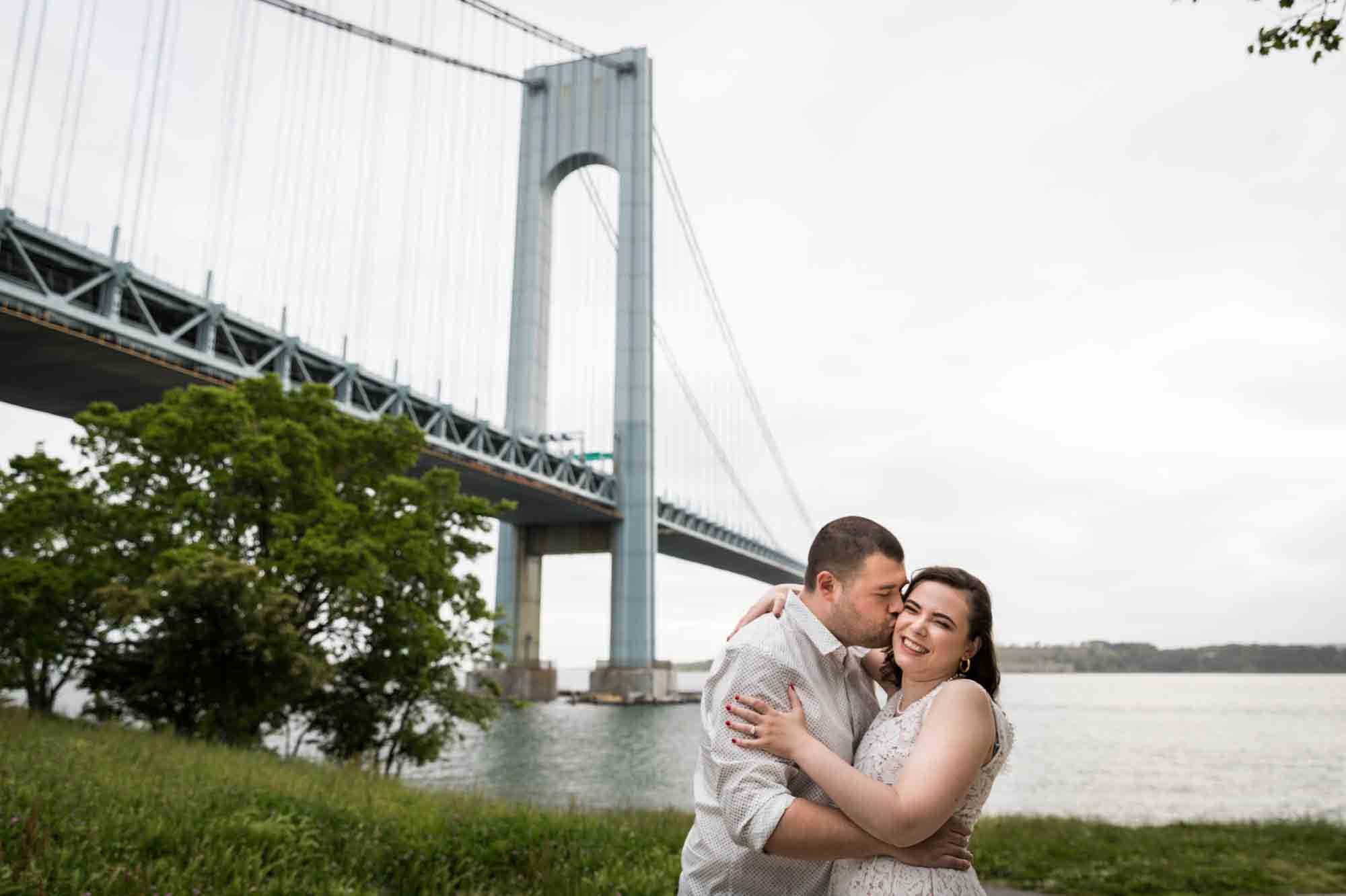 Man kissing woman on the cheek in front of Verrazano Bridge for an article on how to recreate your love story in your engagement shoot