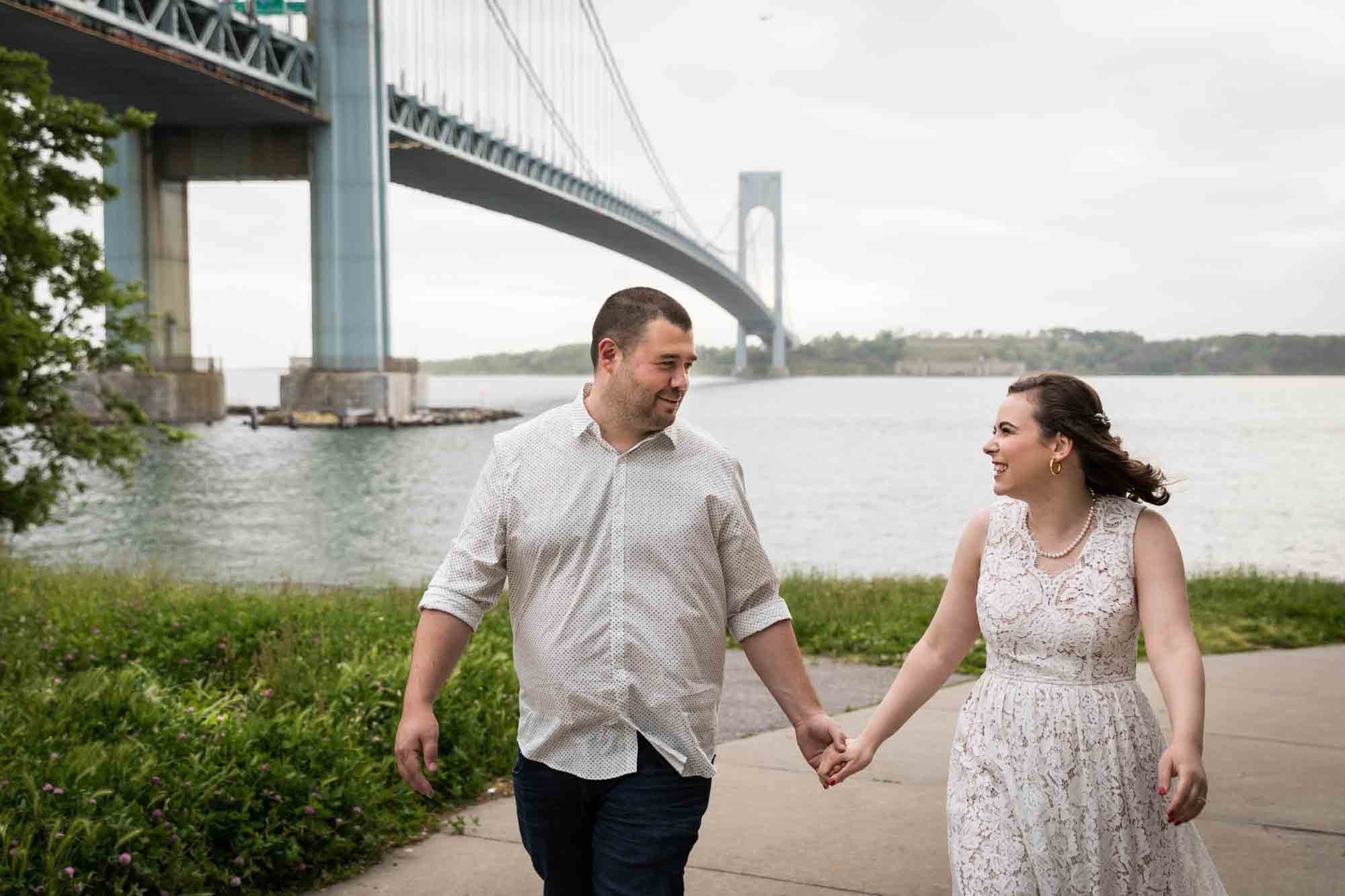 Couple walking on pathway holding hands in front of Verrazano Bridge during a Dyker Heights engagement shoot