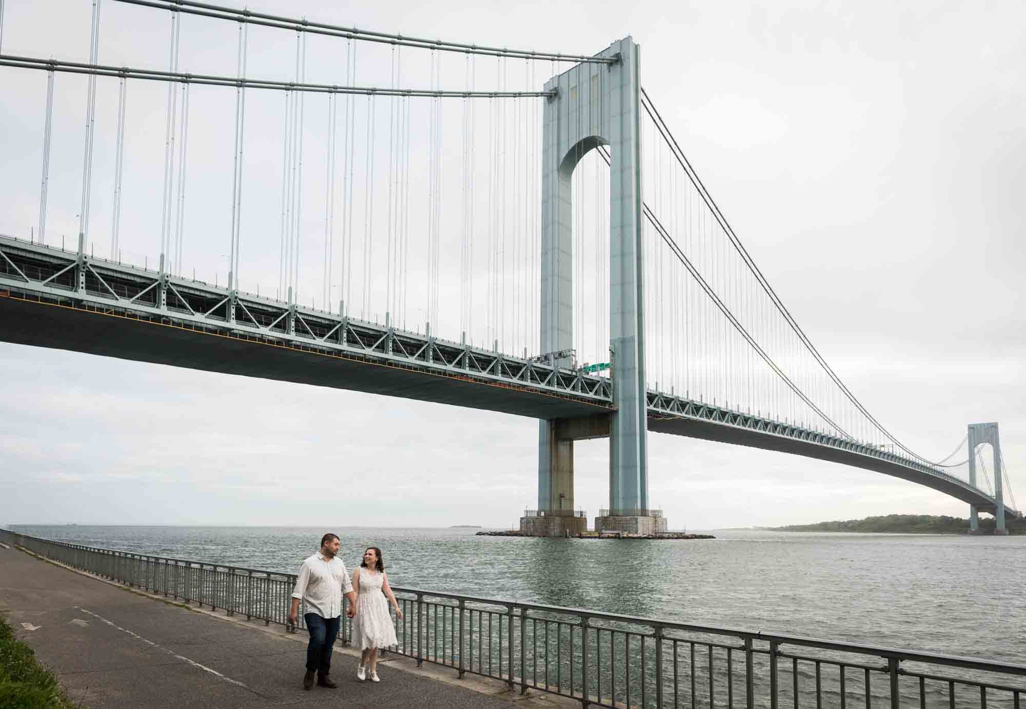 Couple walking on pathway in front of Verrazano Bridge during a Dyker Heights engagement shoot