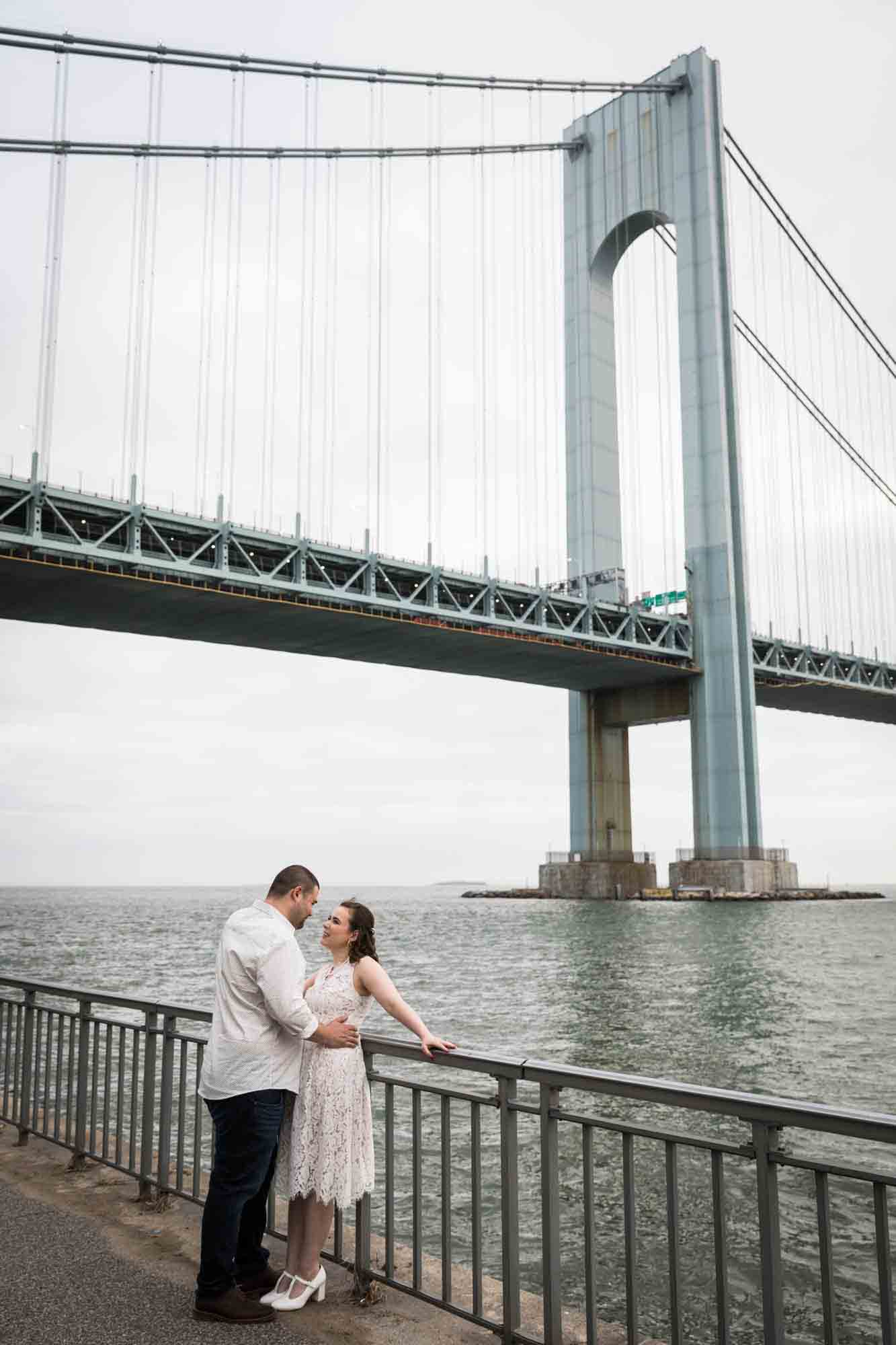 Couple hugging against railing in front of Verrazano Bridge during a Dyker Heights engagement shoot