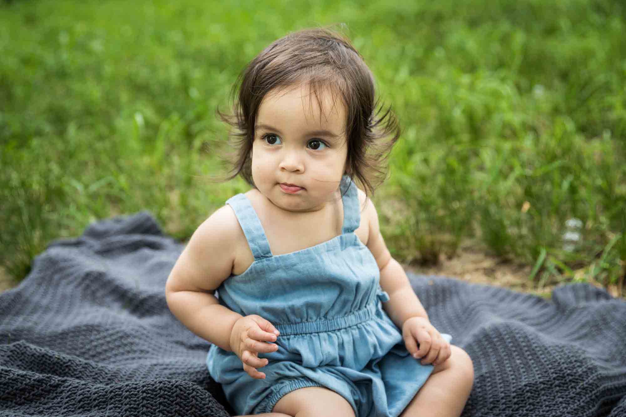 Baby girl wearing blue playsuit and sitting on gray blanket during a Fort Green Park family photo shoot