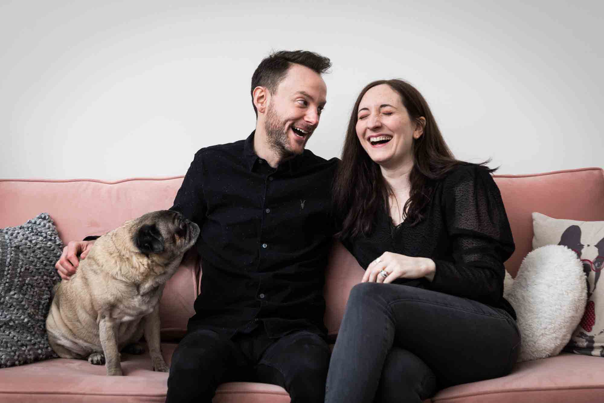 Couple with pug dog sitting on pink couch laughing