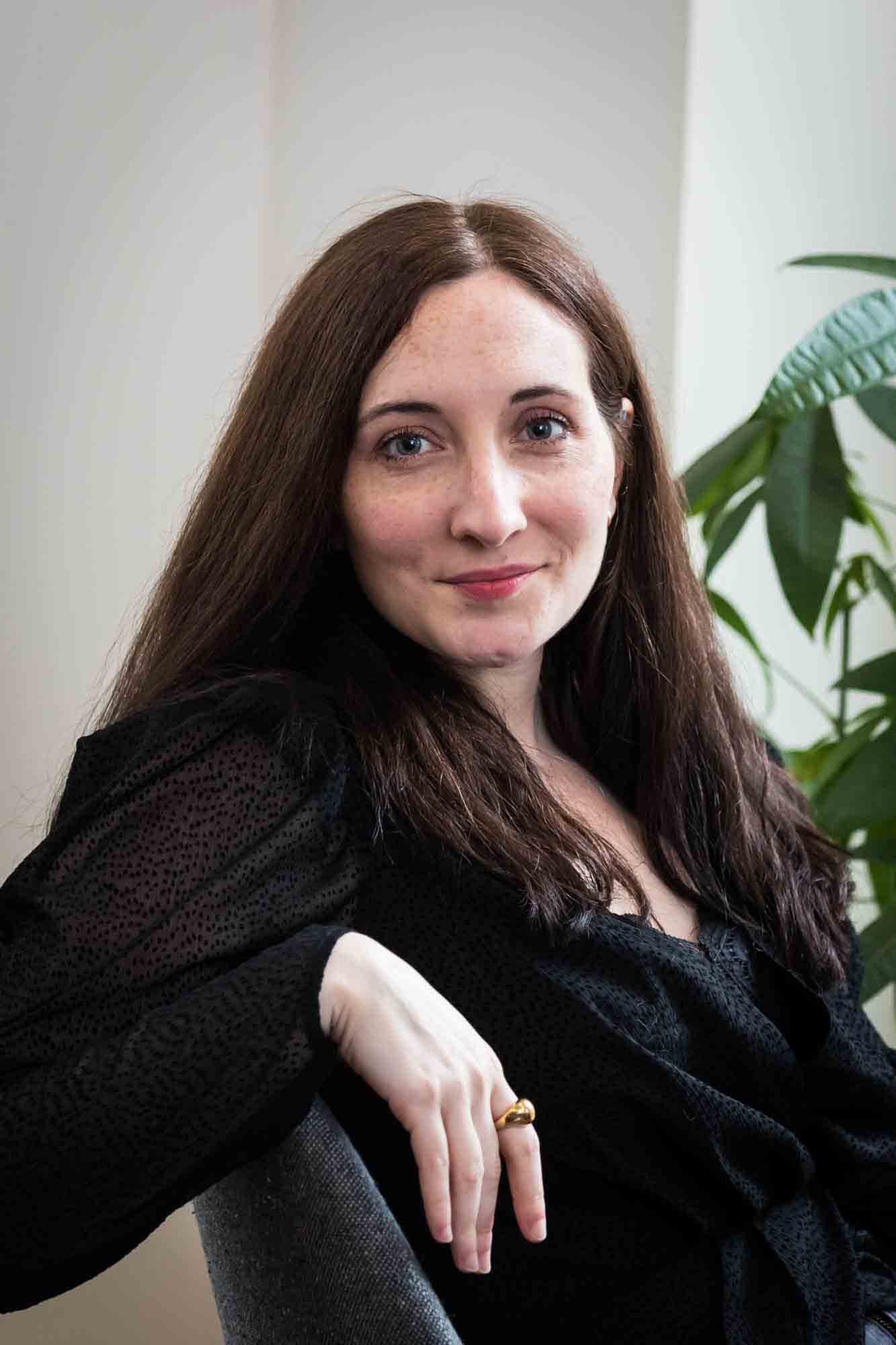 Female author wearing black blouse sitting in chair with plant for an article on professional headshot tips