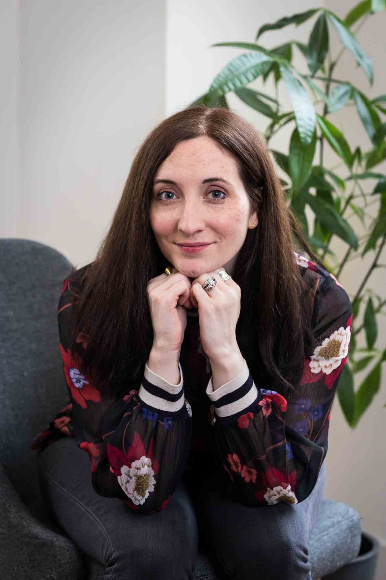 Female author wearing floral blouse sitting in chair with plant for an article on professional headshot tips