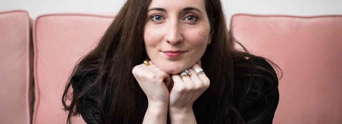 Headshot of female author with hands under chin on pink couch for an article on professional headshot tips