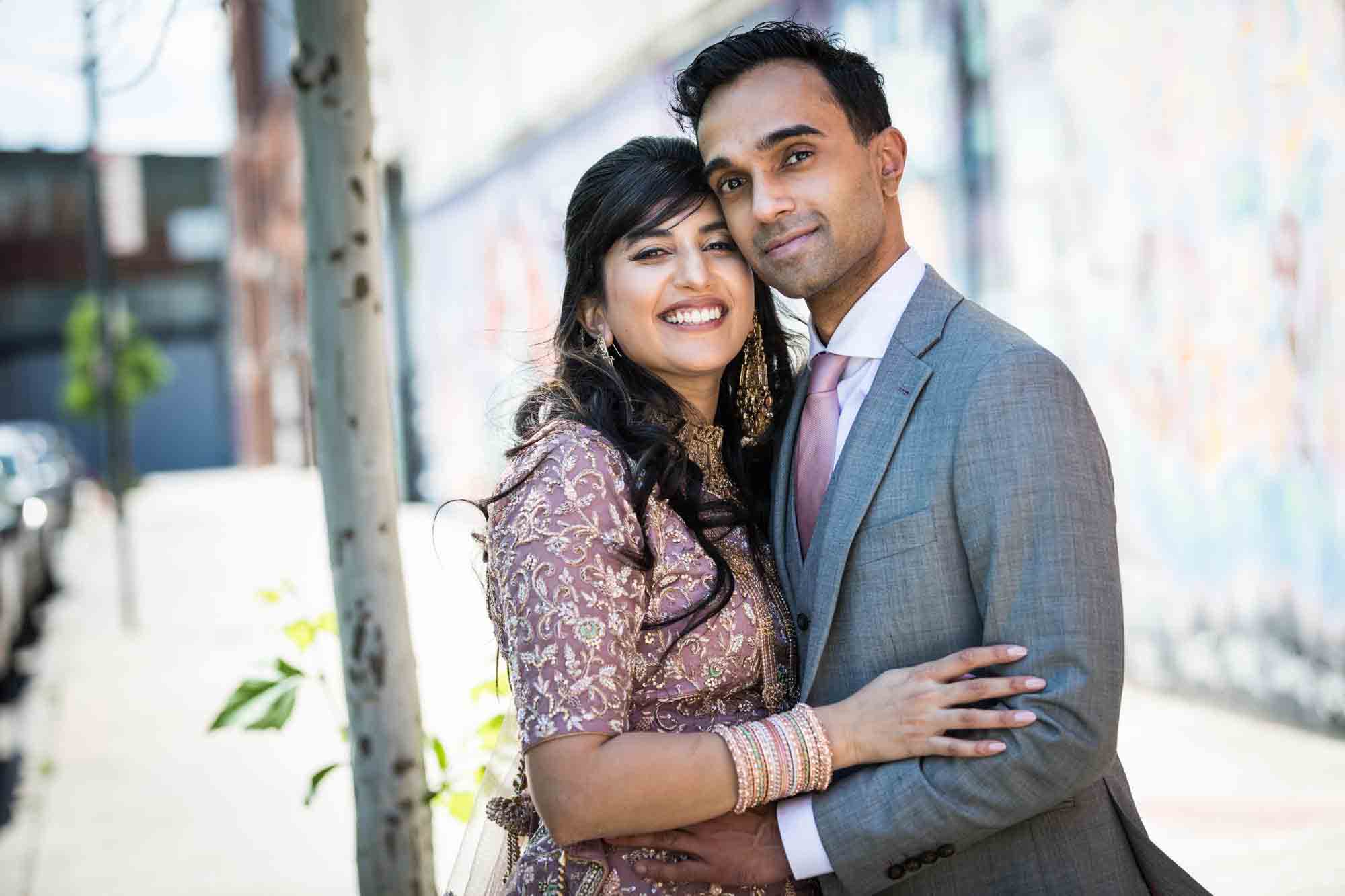 Bride wearing pink Indian wedding dress and groom in grey suit hugging for article on wedding reception game ideas