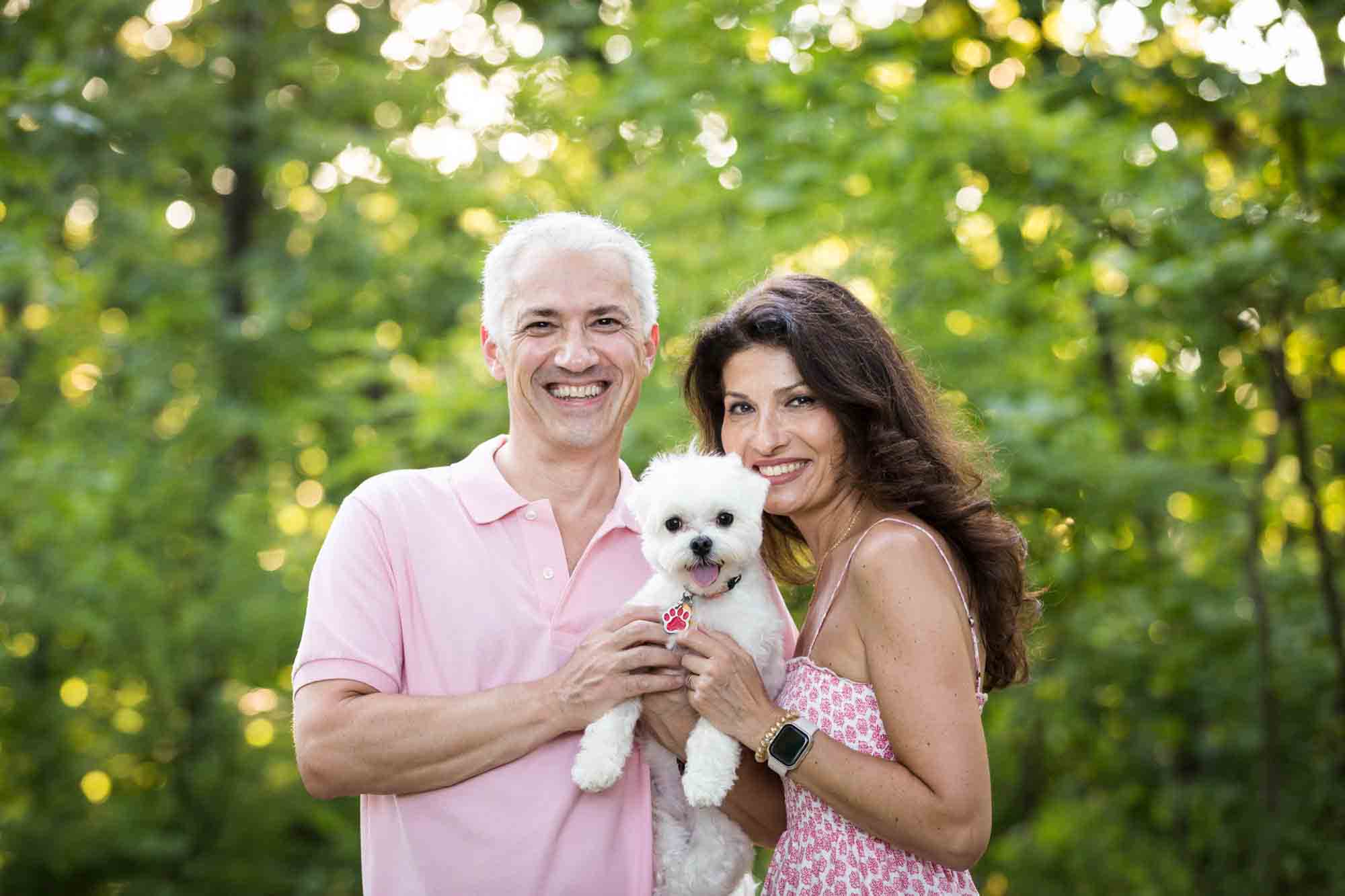 Couple holding small, white dog in front of trees during a Forest Park family photo shoot