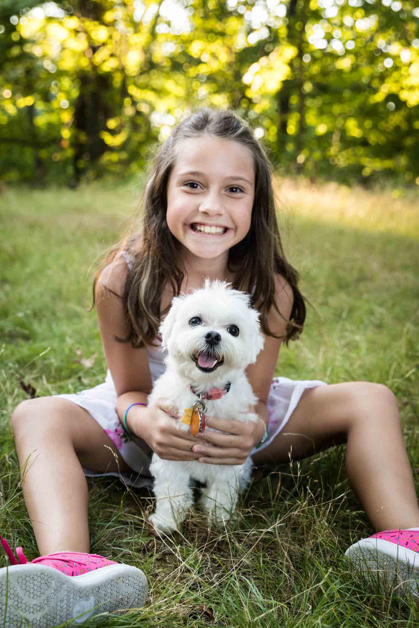 Young girl with long brown hair sitting on ground holding small, white dog