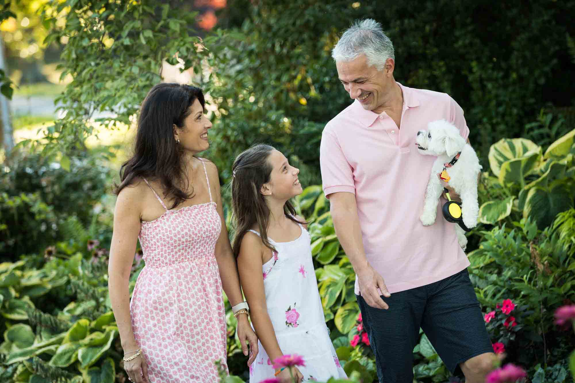 Parents holding small white dog and young girl walking in garden during a Forest Park family photo shoot