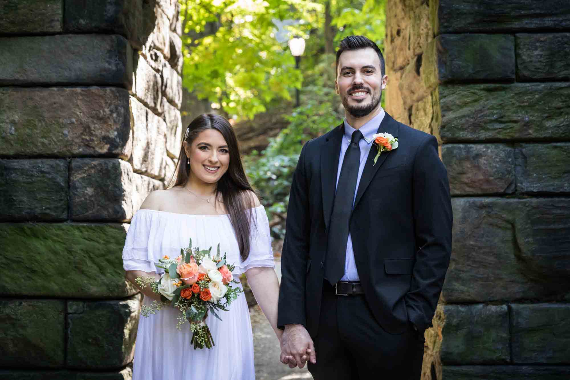 Bride and groom walking hand-in-hand under stone arch Article on how to elope in Central Park by NYC wedding photojournalist, Kelly Williams. Includes photos from a real wedding near Bow Bridge.