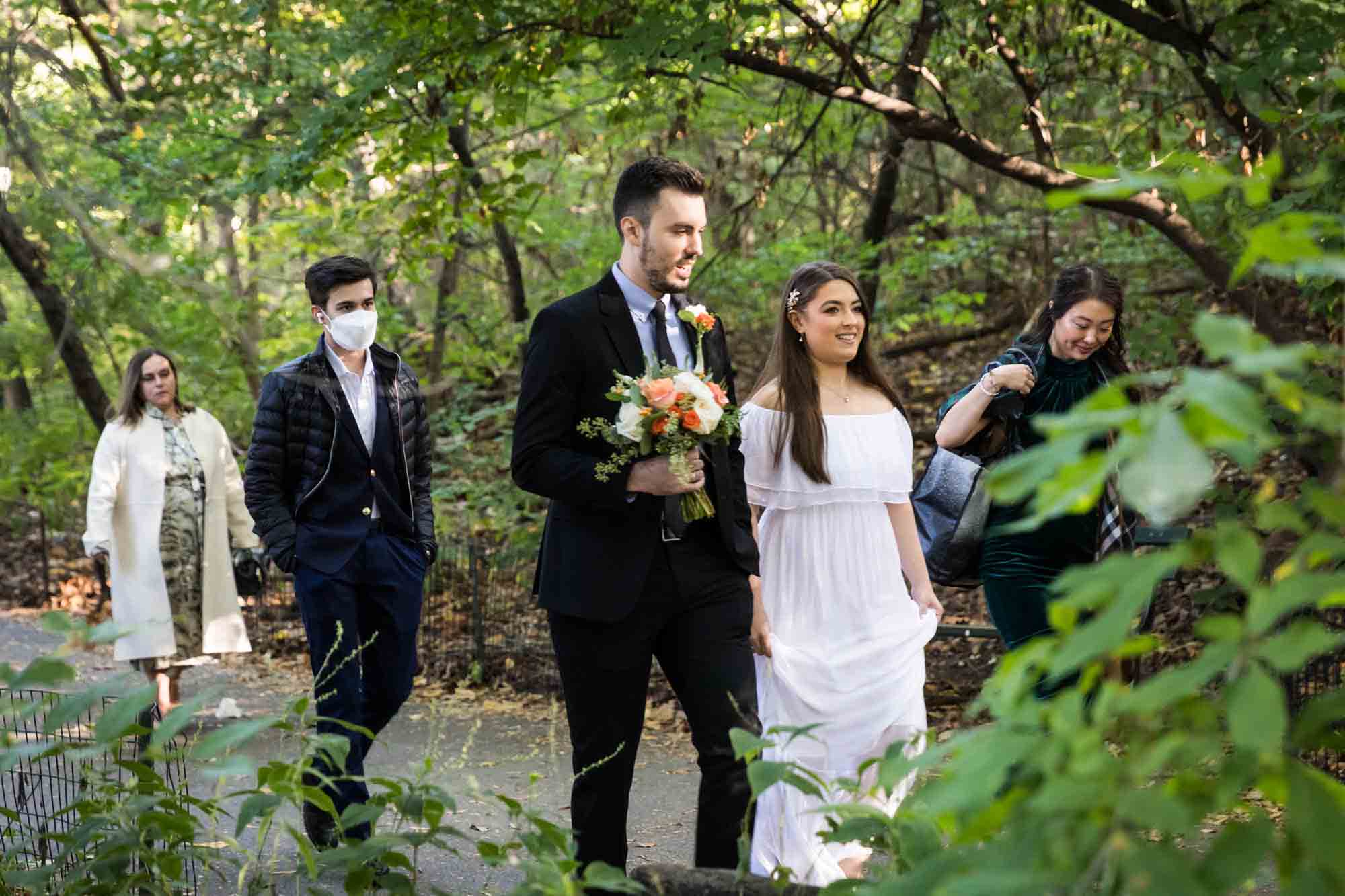 Group walking in forest with groom holding flower bouquet for article on how to elope in Central Park