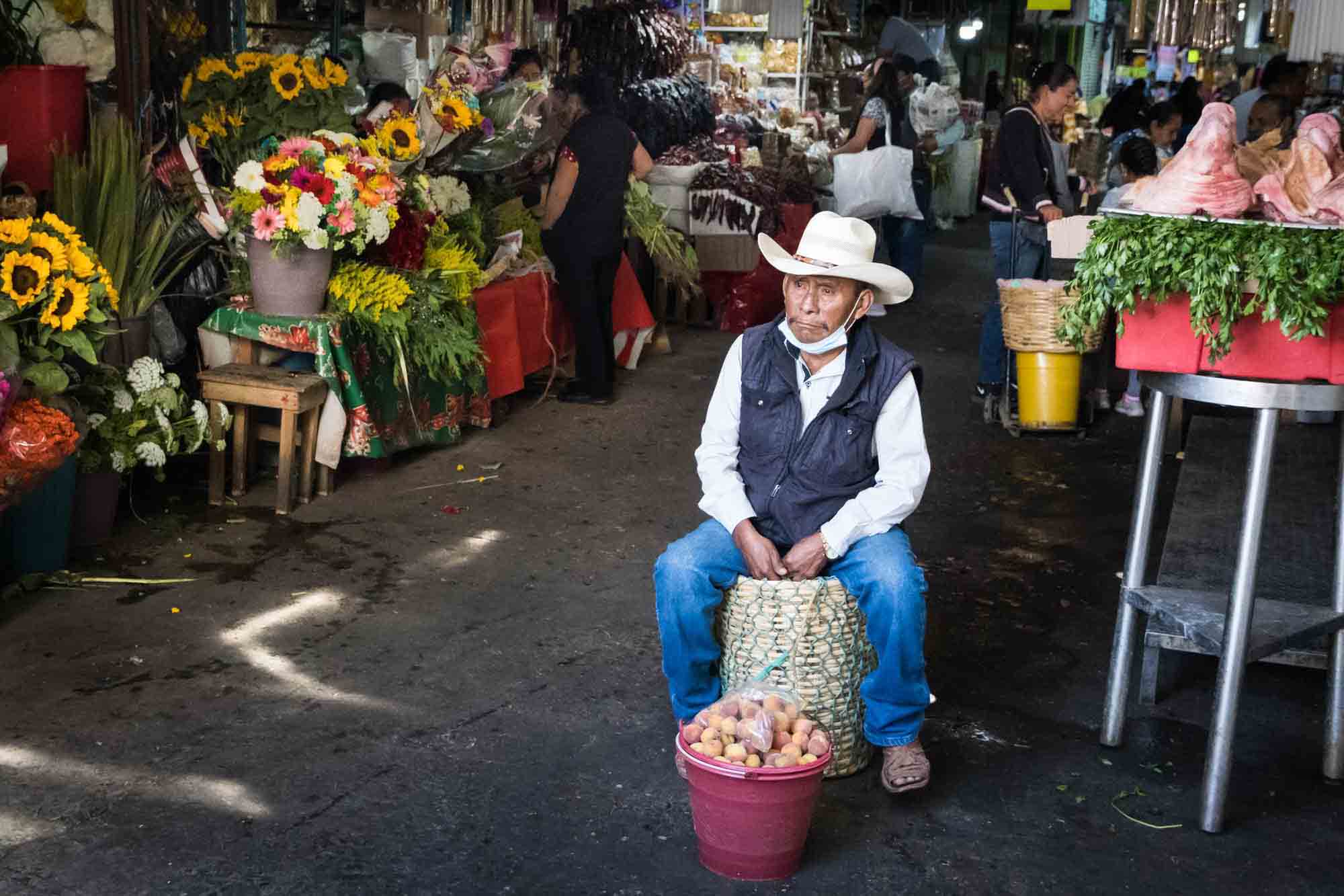 A man wearing a cowboy hat sits at the Oaxaca market selling a bucket of peaches