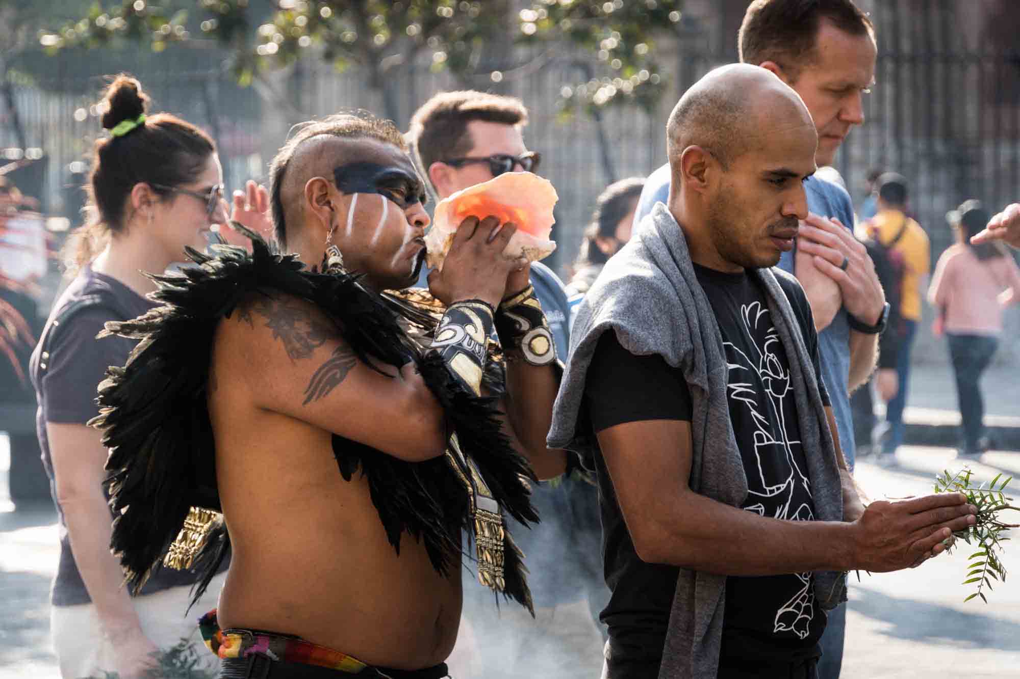 An indigenous man blows a shell behind another man in a Mexico City ceremony