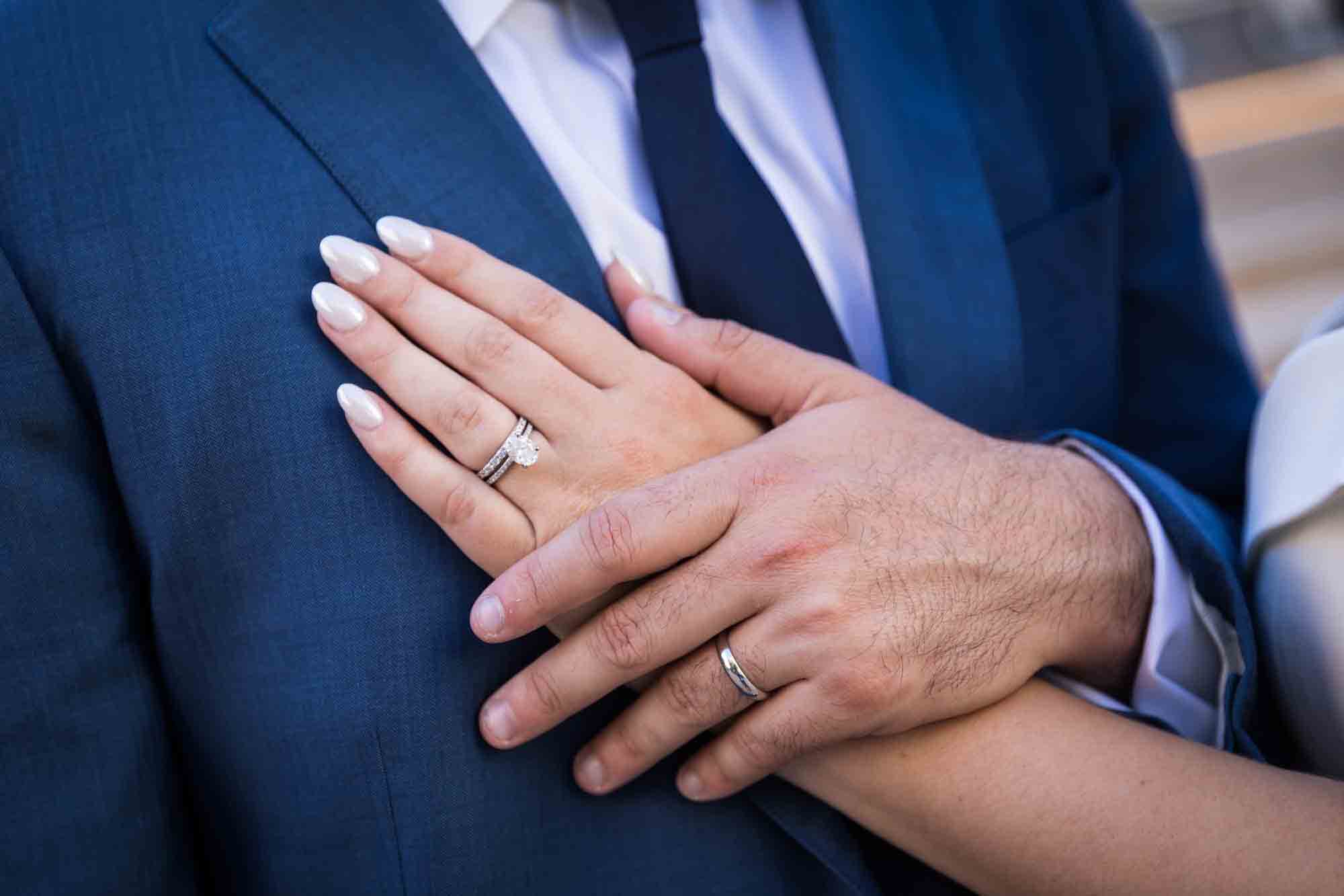 Close up of bride and groom's hands wearing wedding rings held on man's blue jacket