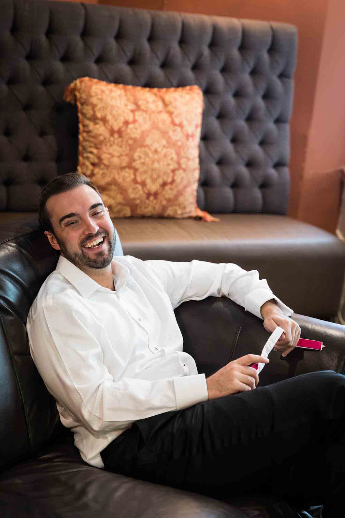 Groom laughing while wearing white shirt and sitting on leather couch at a Briarcliff Manor wedding