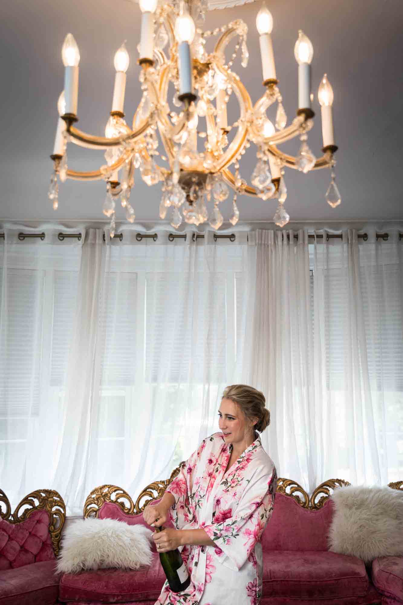 Bride wearing floral robe opening bottle of champagne under chandelier and in front of pink couch at a Briarcliff Manor wedding