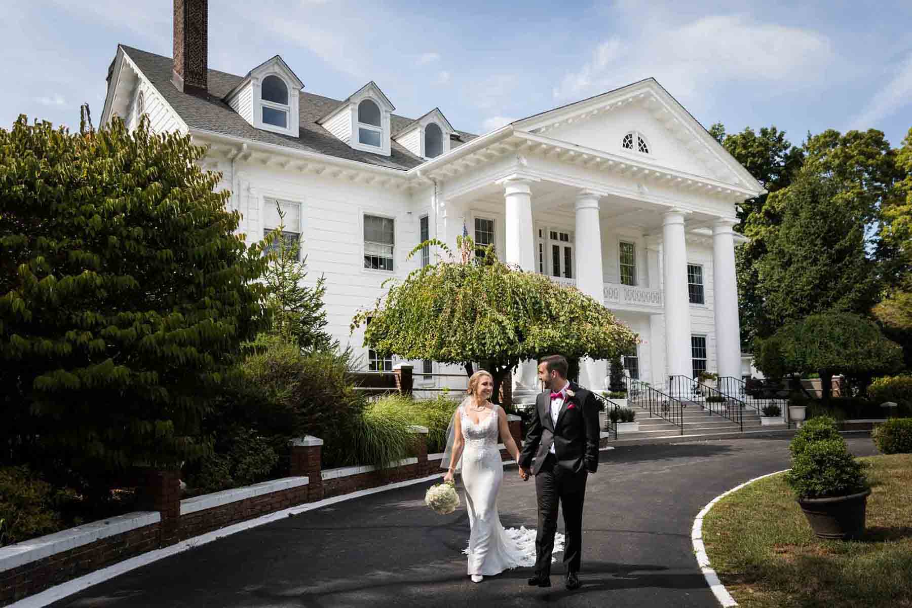 Bride and groom holding hands and walking in front of historical white home with Greek columns at a Briarcliff Manor wedding