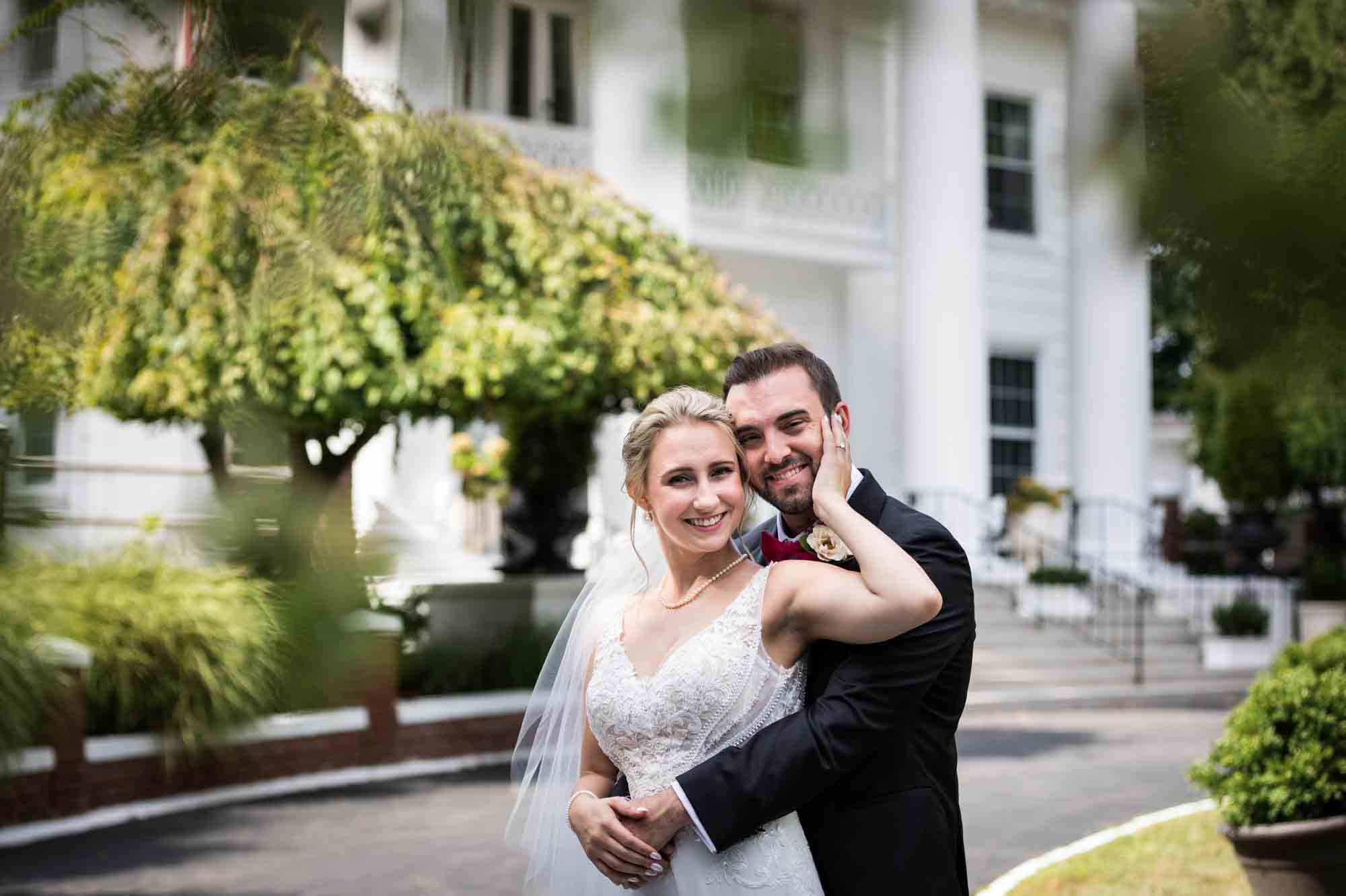 Bride hugging groom with hand on his face in front of white historical home at a Briarcliff Manor wedding