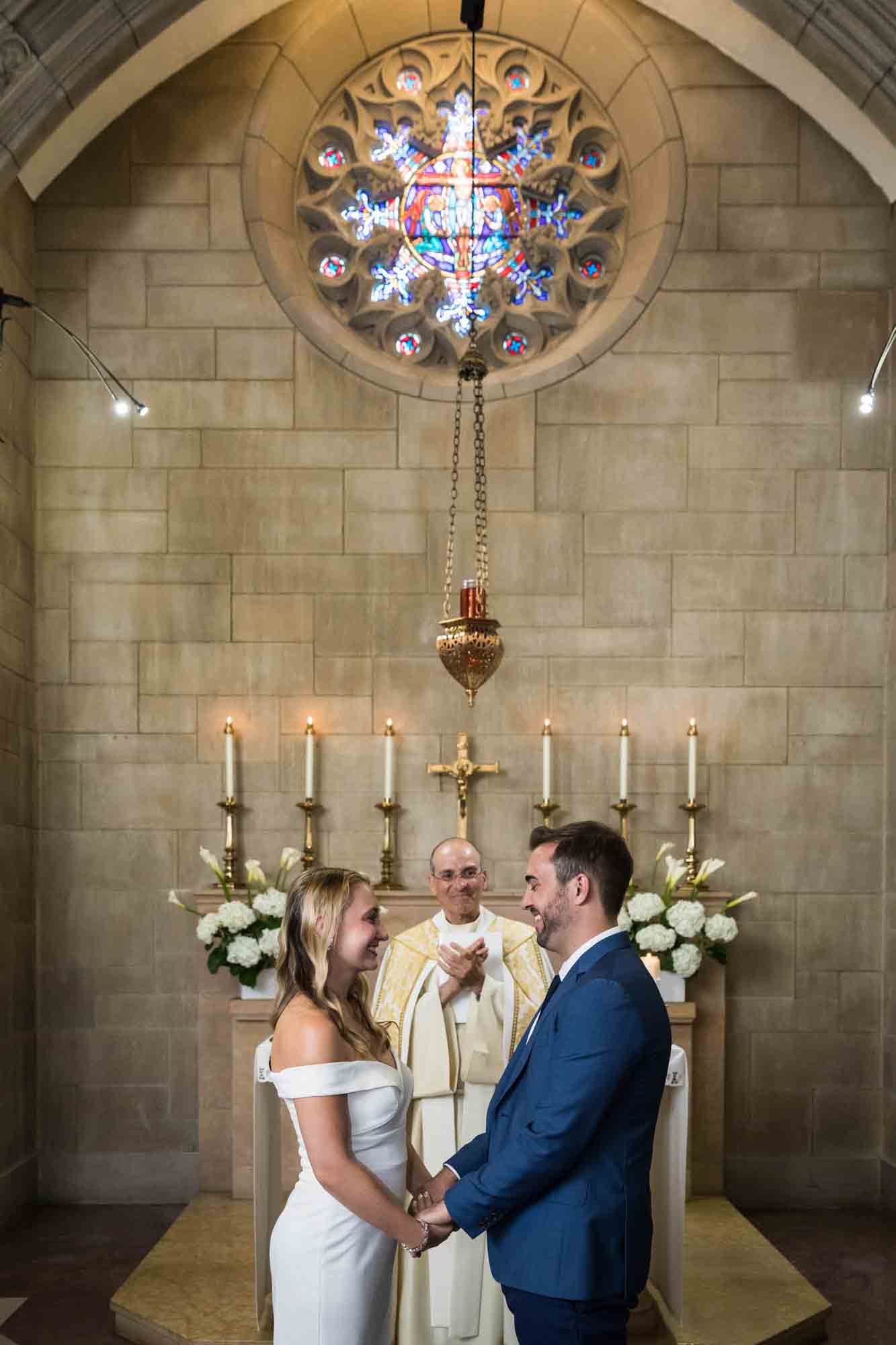Bride and groom saying vows in front of priest in stone chapel at St. Paul the Apostle Church