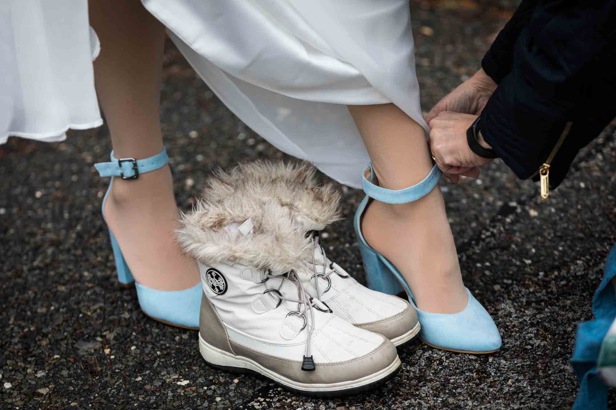 Close up of woman's hands adjusting strap on bride's blue high heels with white snow boots in frame