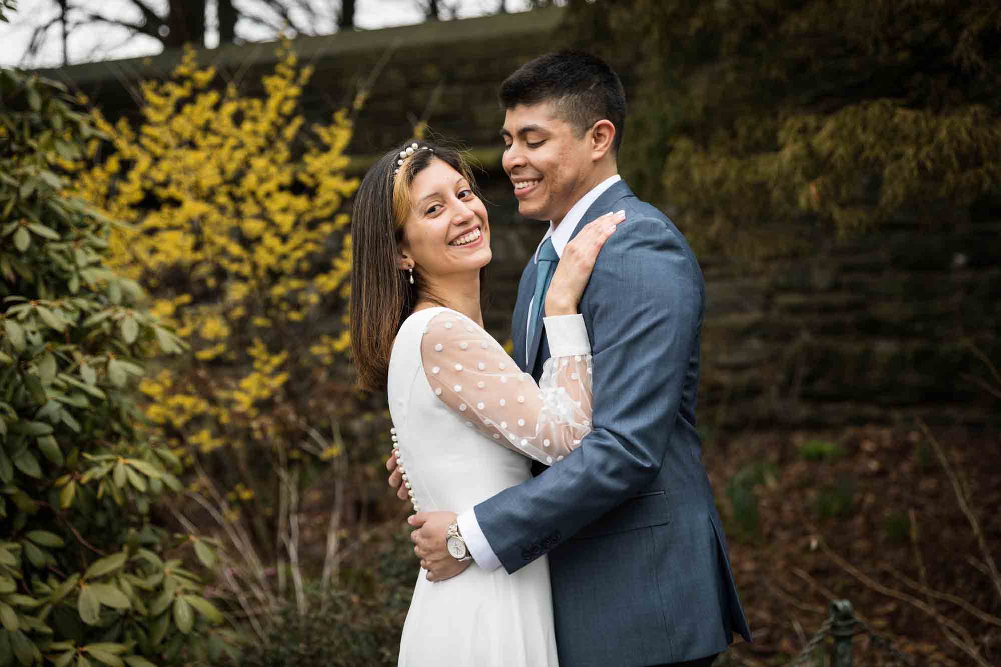 Bride and groom hug in front of yellow forsythia tree in Fort Tryon Park Heather Garden