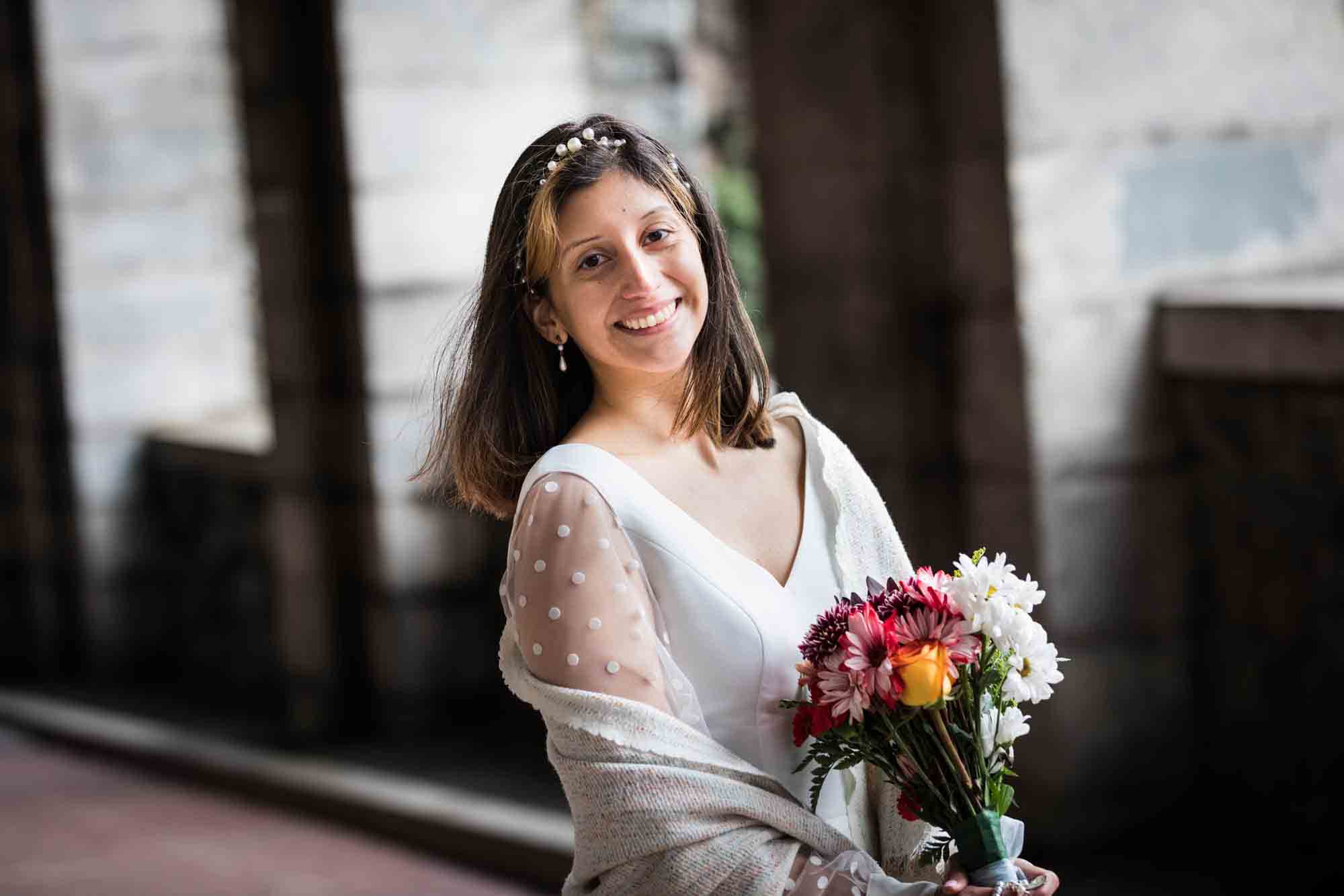Bride wearing white shawl and holding colorful flowers under Billings Arcade in Fort Tryon Park
