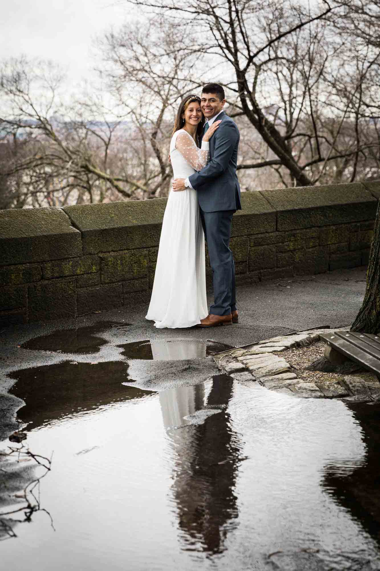 A bride and groom hug in front of water puddle during a Fort Tryon Park wedding