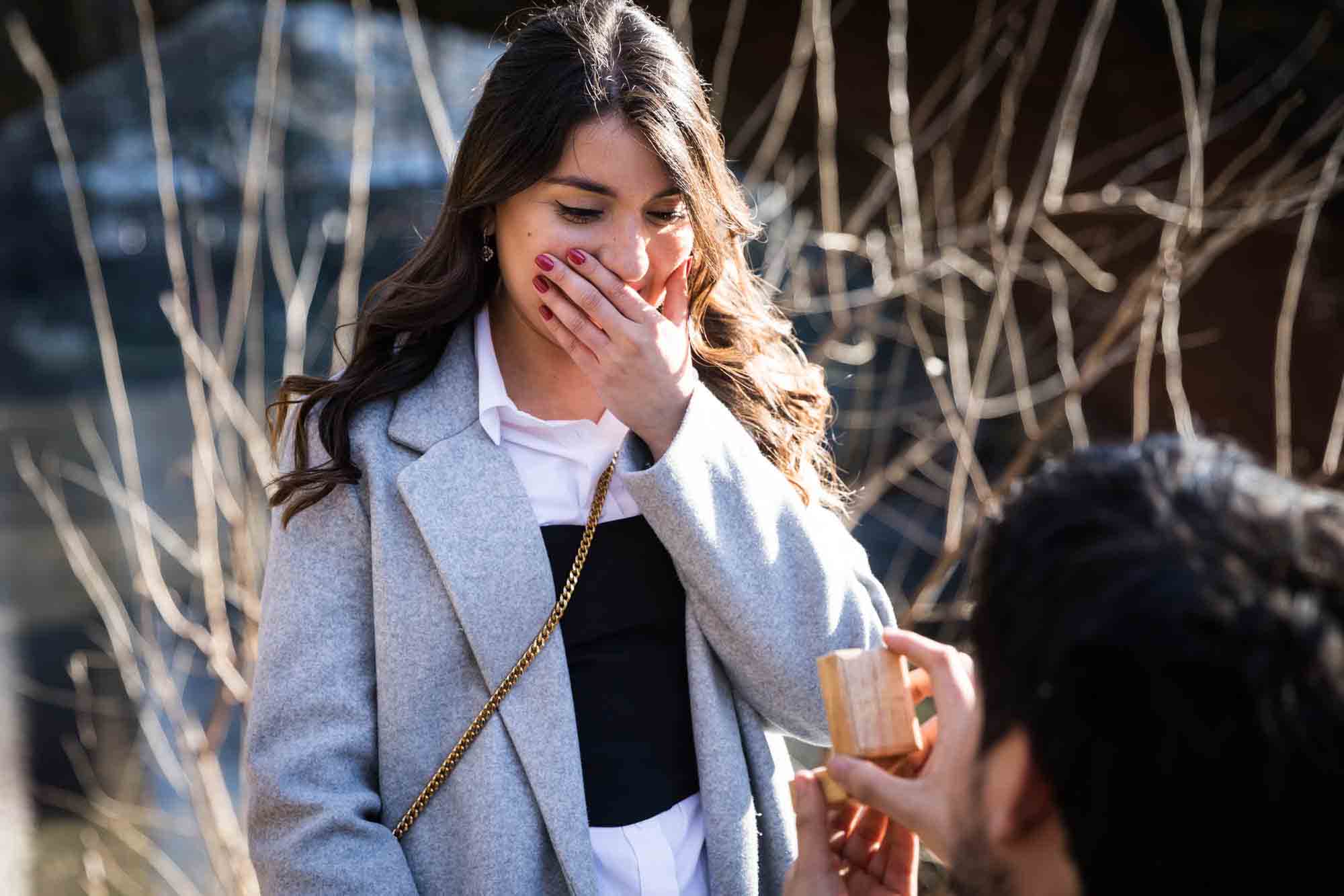 Woman with hand over mouth looking down at man holding engagement ring box during a Gapstow Bridge surprise proposal in Central Park