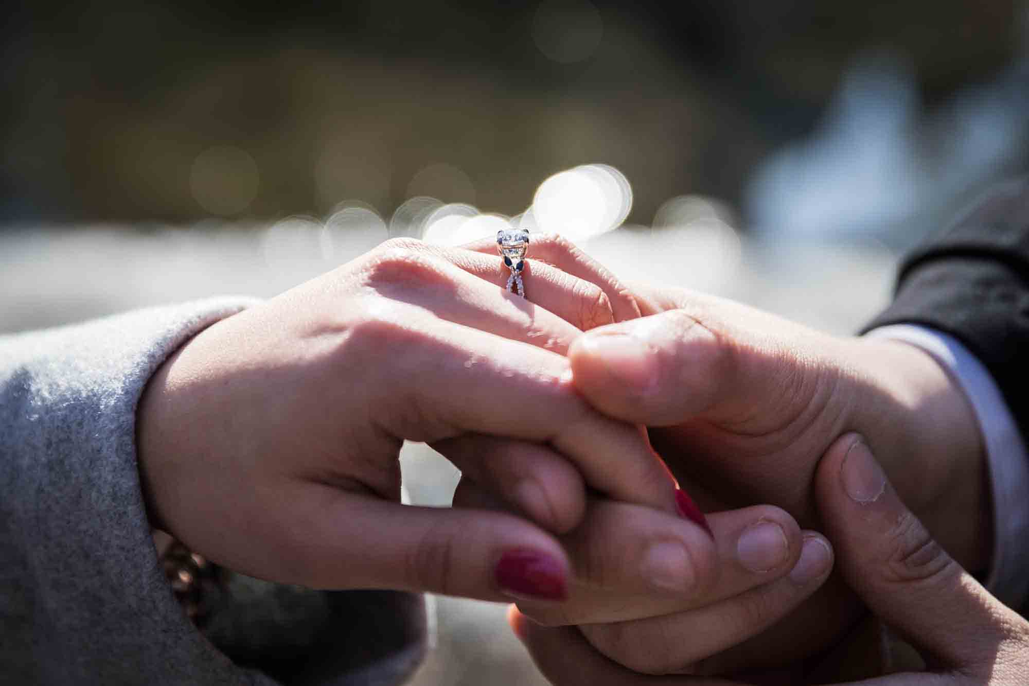 Close up of man holding woman's hand wearing wearing diamond engagement ring during a Gapstow Bridge surprise proposal in Central Park