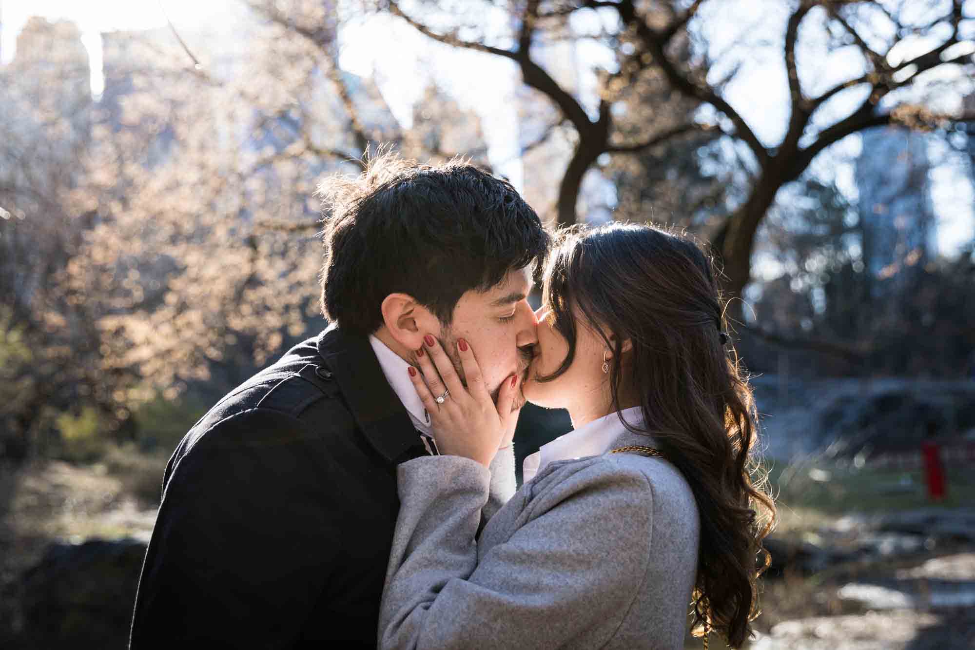 Man and woman kissing with woman's hand showing engagement ring during a Gapstow Bridge surprise proposal in Central Park