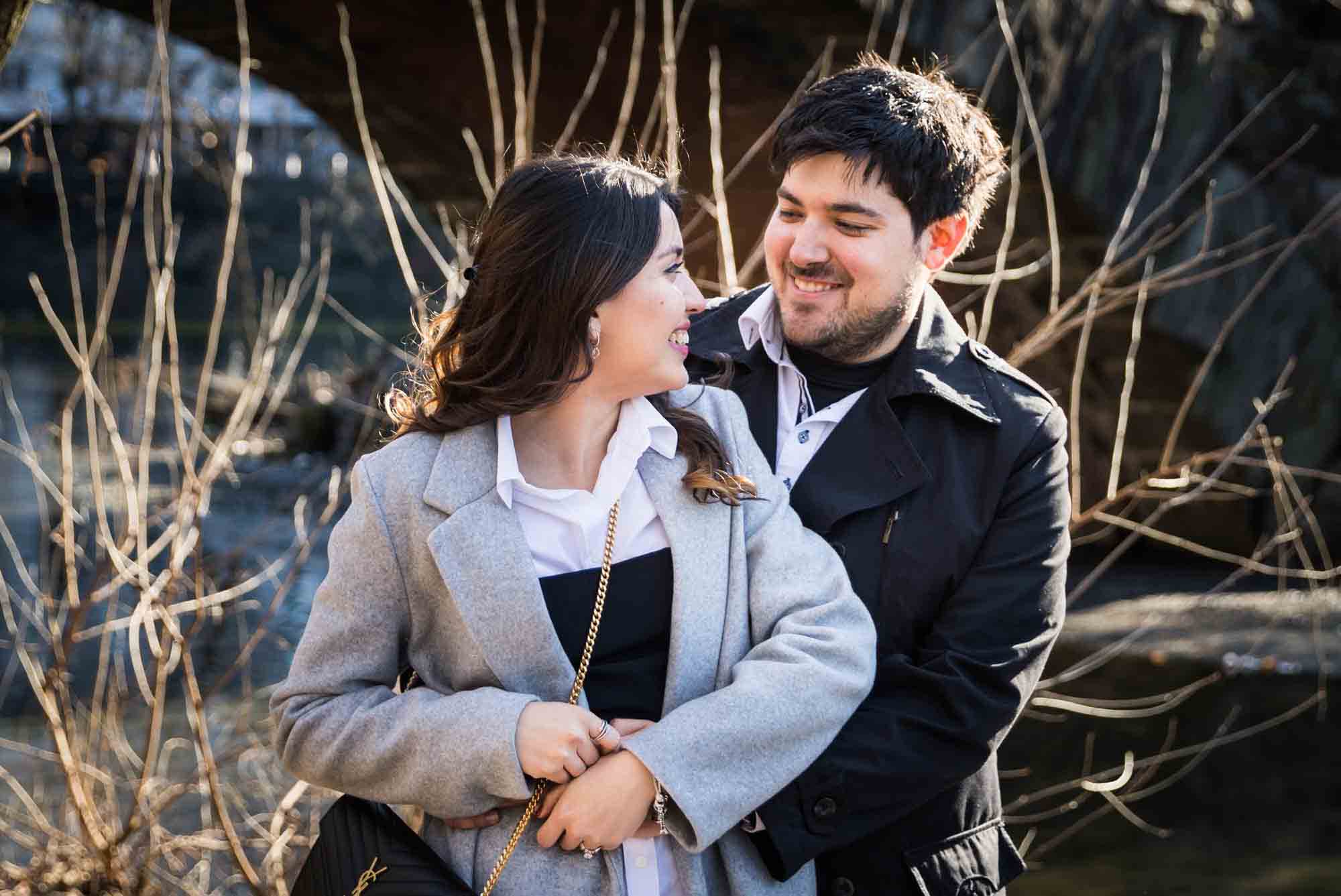 Couple hugging with man behind woman during a Gapstow Bridge surprise proposal in Central Park