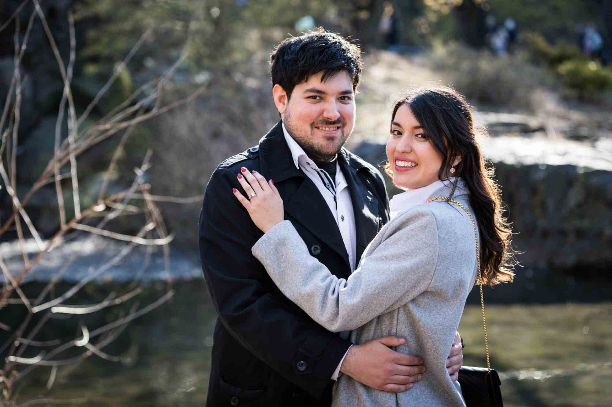 Couple hugging with woman's hand on man's shoulder during a Gapstow Bridge surprise proposal in Central Park