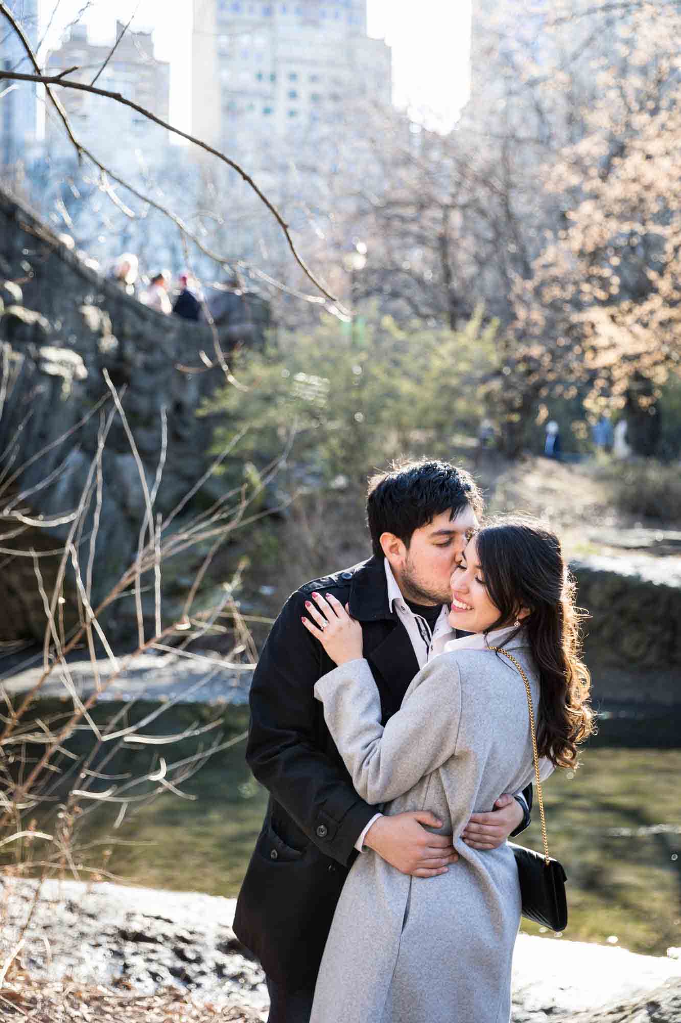 Man kissing woman on the cheek in front of bridge during a Central Park surprise proposal