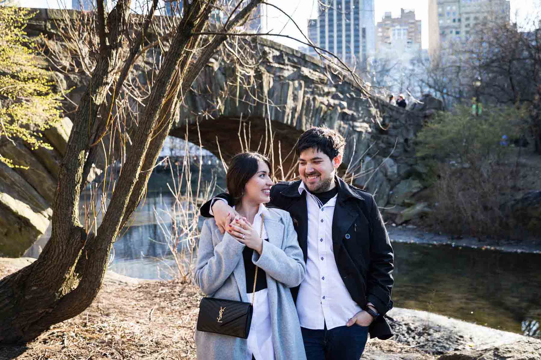 Couple walking in front of bridge with man's arm around woman during a Central Park surprise proposal