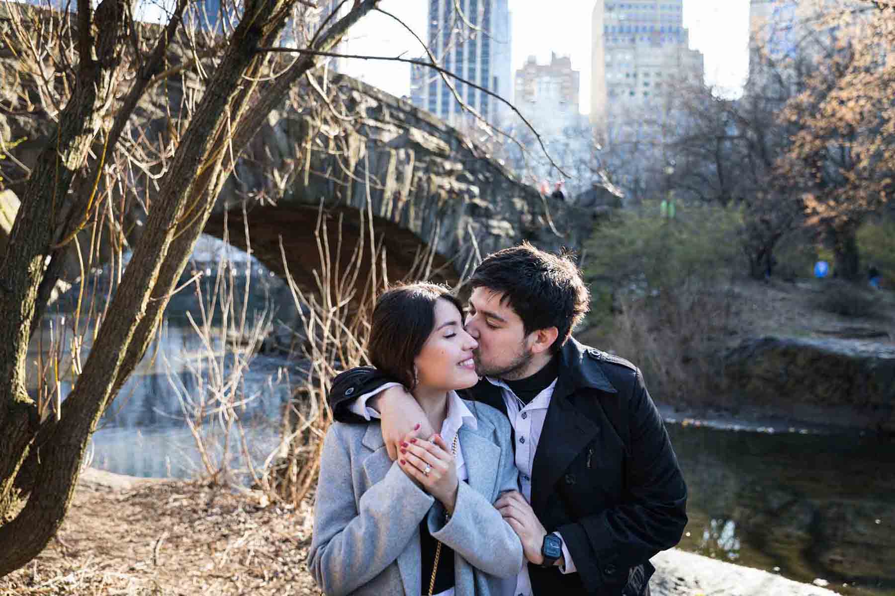 Man kissing woman on the cheek in front of bridge during a Central Park surprise proposal