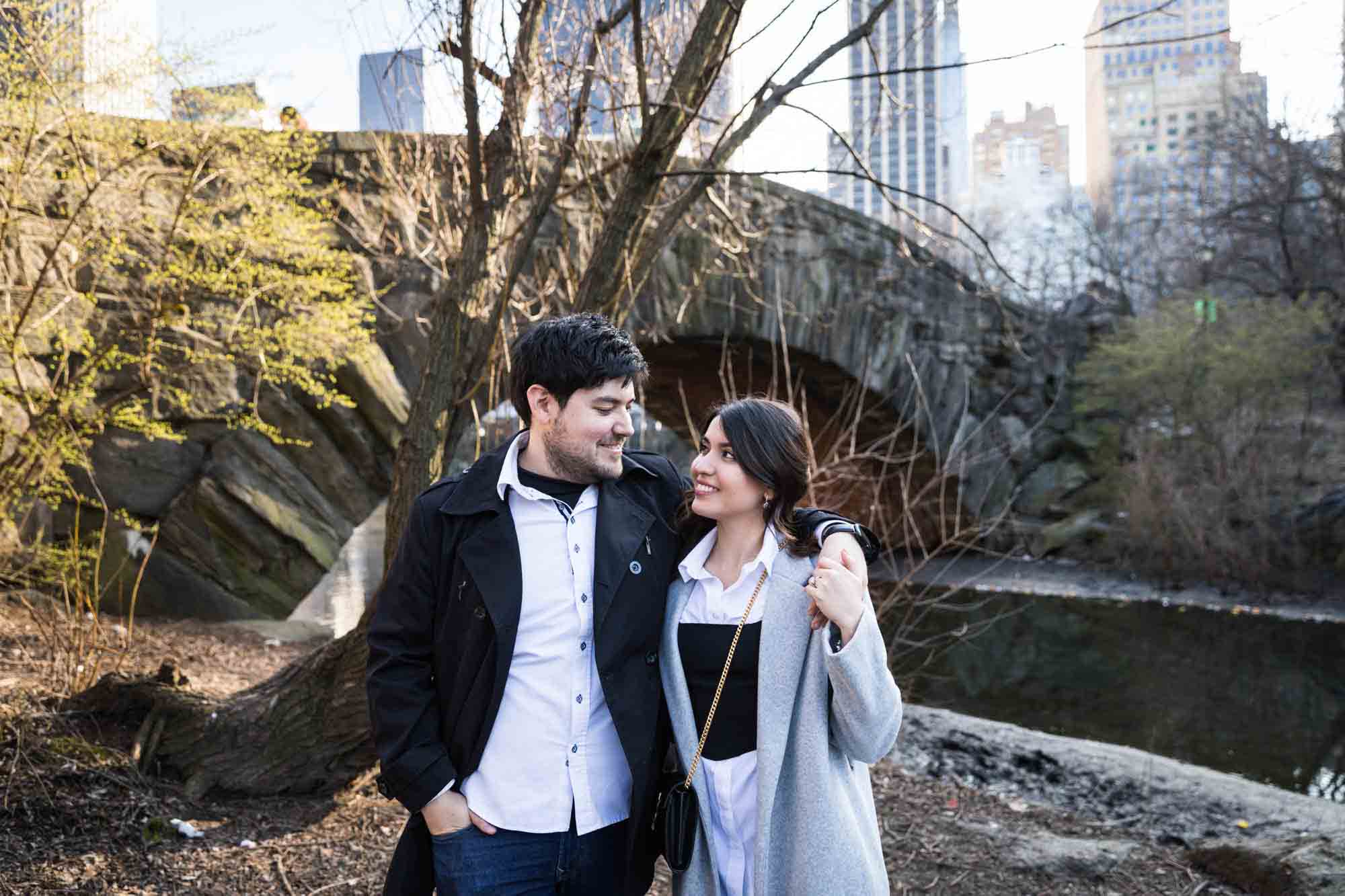 Couple walking in front of bridge with man's arm around woman during a Central Park surprise proposal