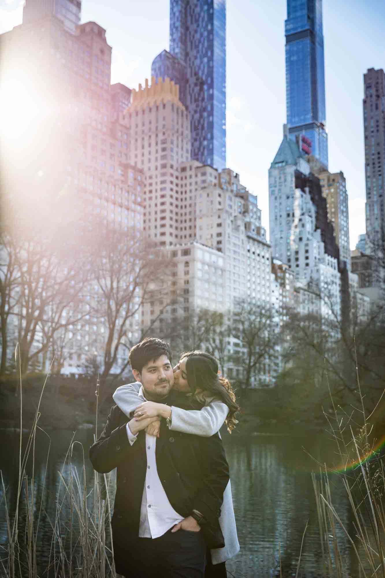 Woman kissing man on the cheek in front of Central Park lake and NYC skyline during a Central Park surprise proposal
