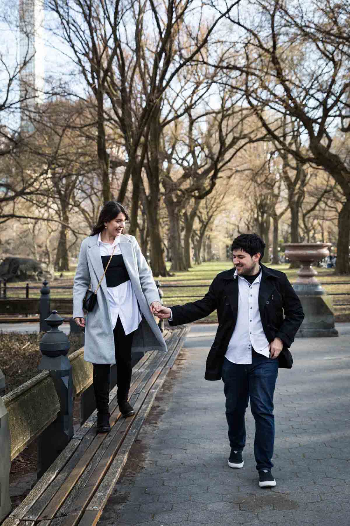 Woman walking on benches while holding man's hand in Central Park after a NYC surprise proposal