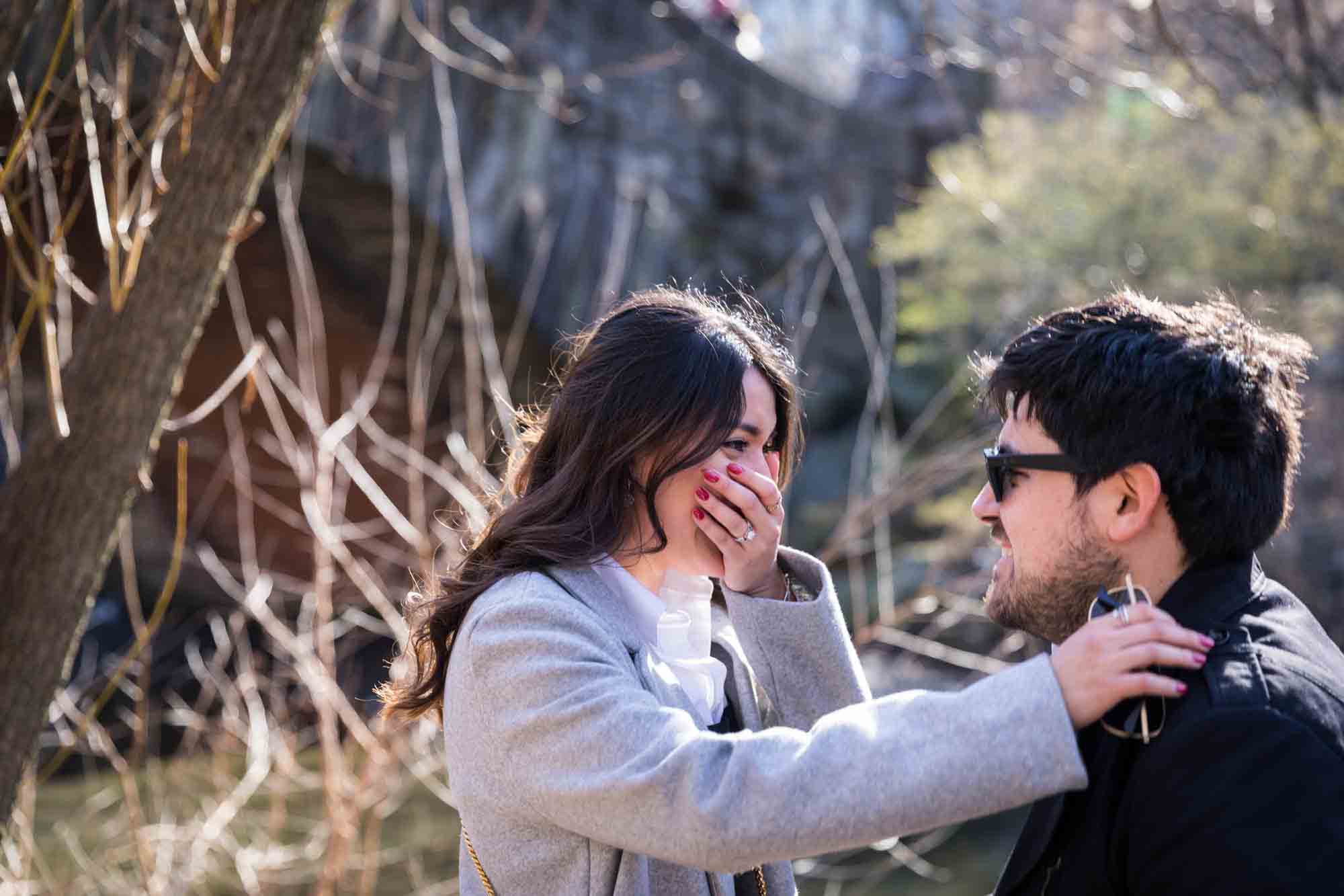 Surprised woman with hand over mouth looking at man wearing sunglasses during a Gapstow Bridge surprise proposal in Central Park