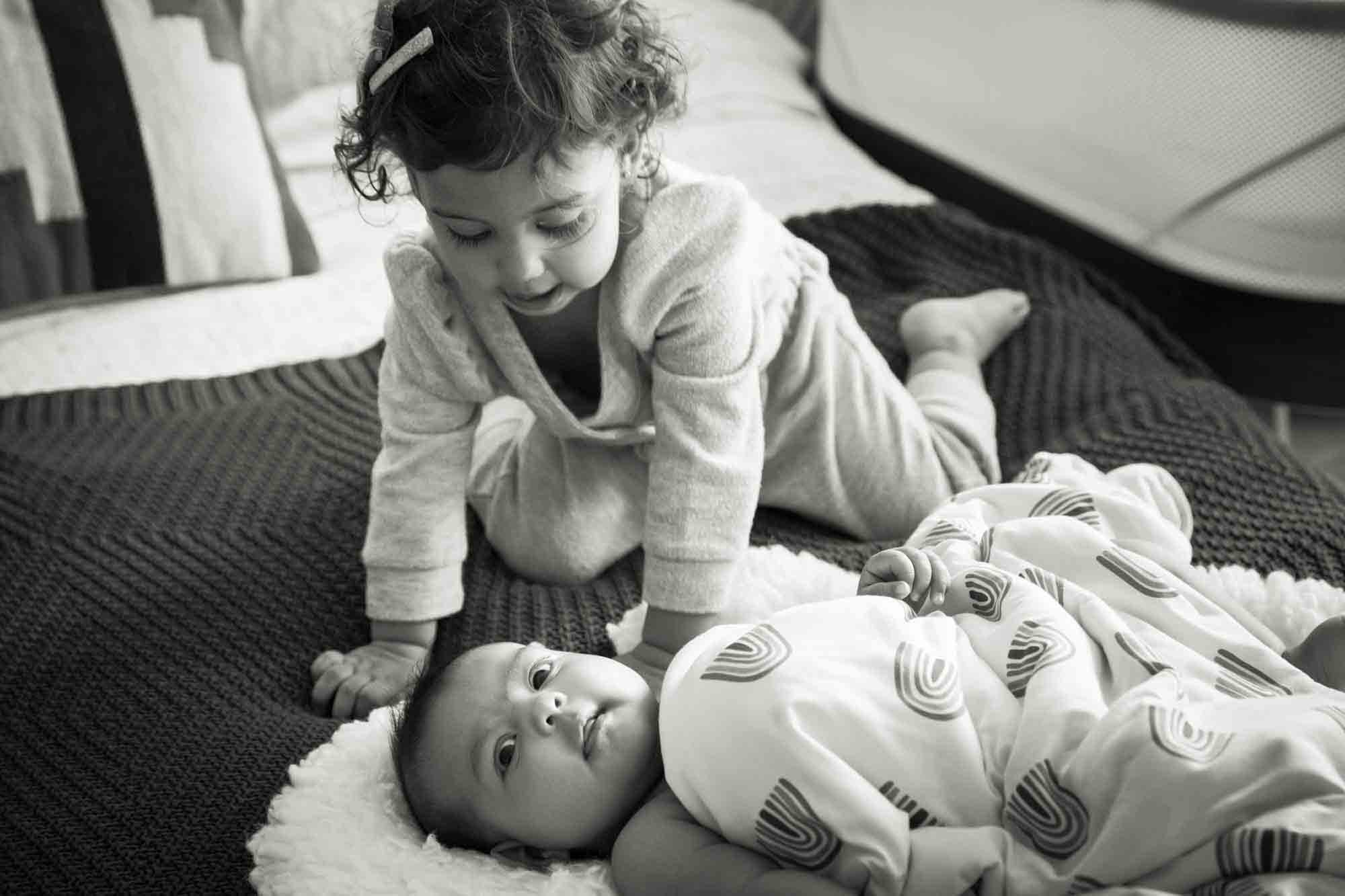 Black and white photo of toddler looking down on baby lying on bed