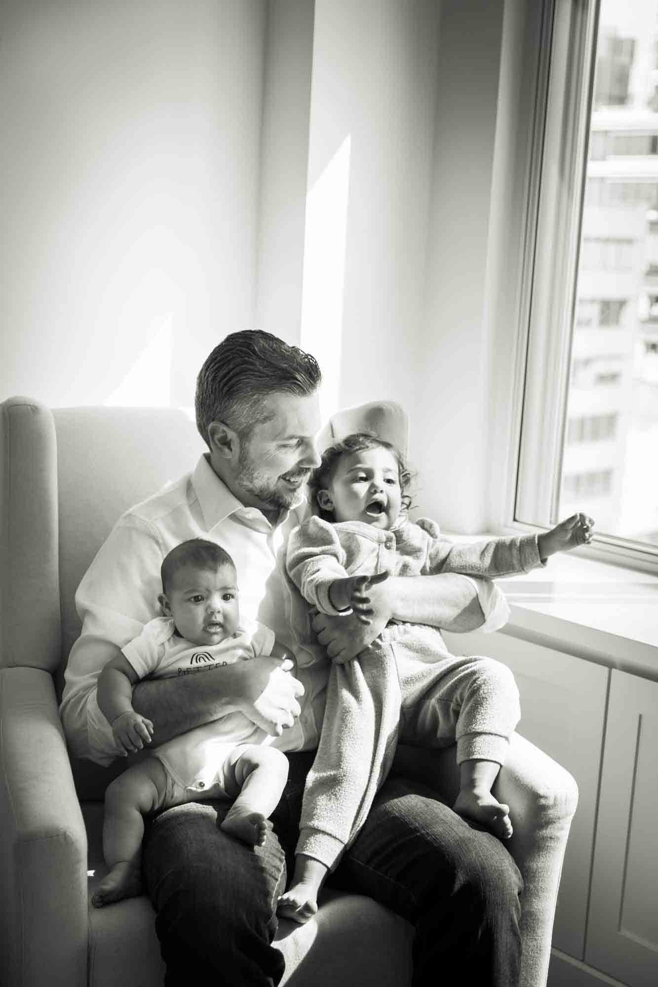 Black and white photo of father sitting in chair holding toddler and baby by window during a NYC family portrait session
