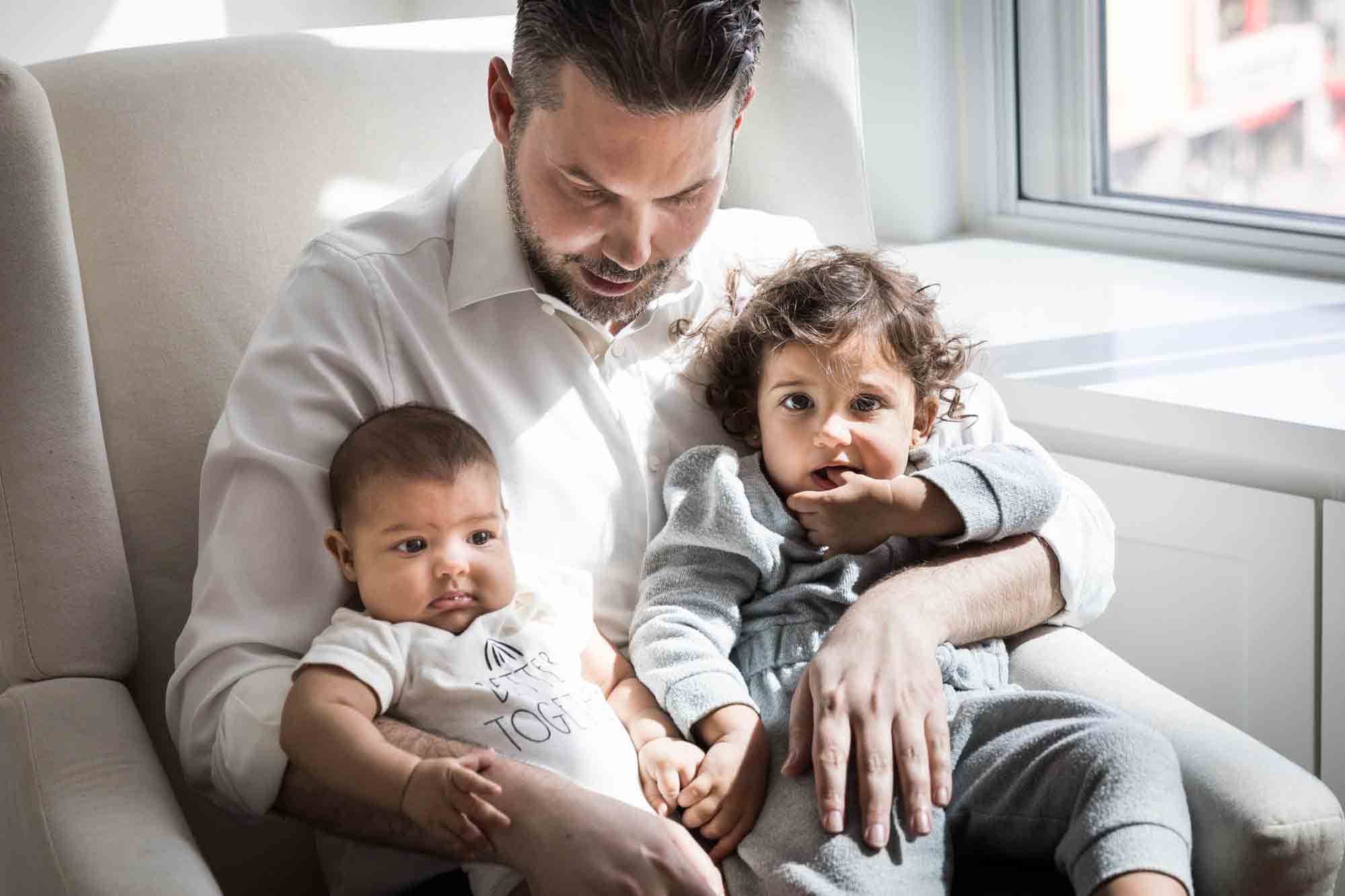 Father sitting in chair holding toddler and baby by window during a NYC family portrait session
