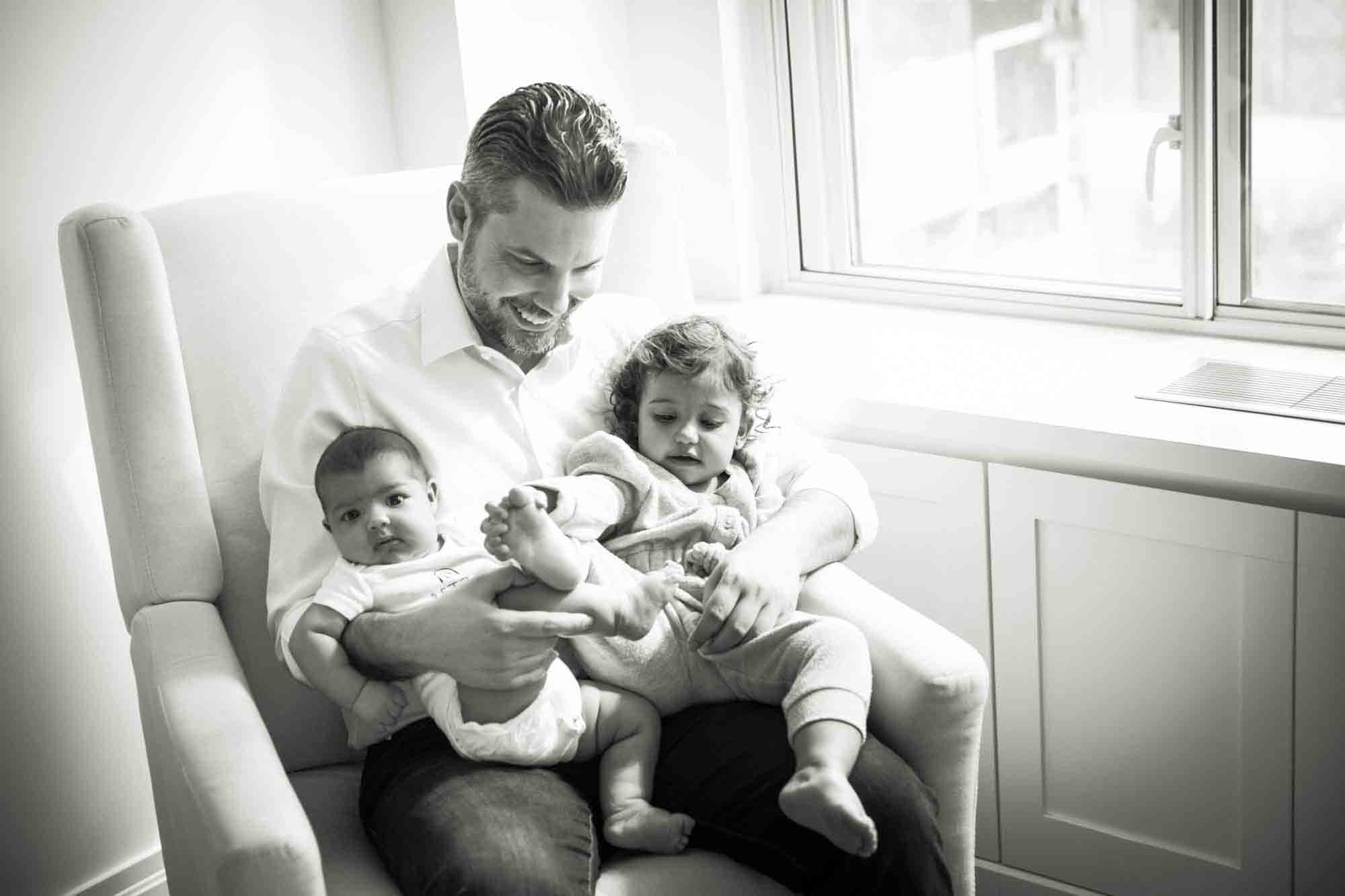 Black and white photo of father sitting in chair holding toddler and baby by window during a NYC family portrait session