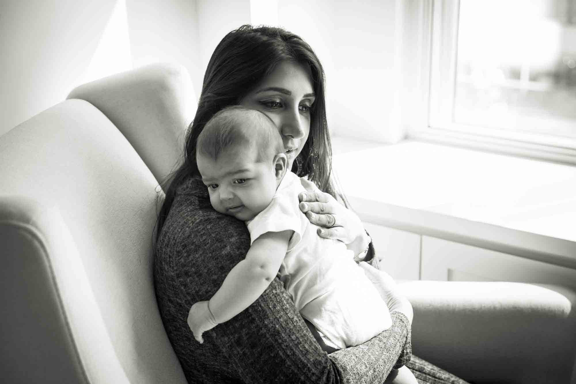 Black and white photo of mother sitting in chair holding baby by window during a NYC family portrait session