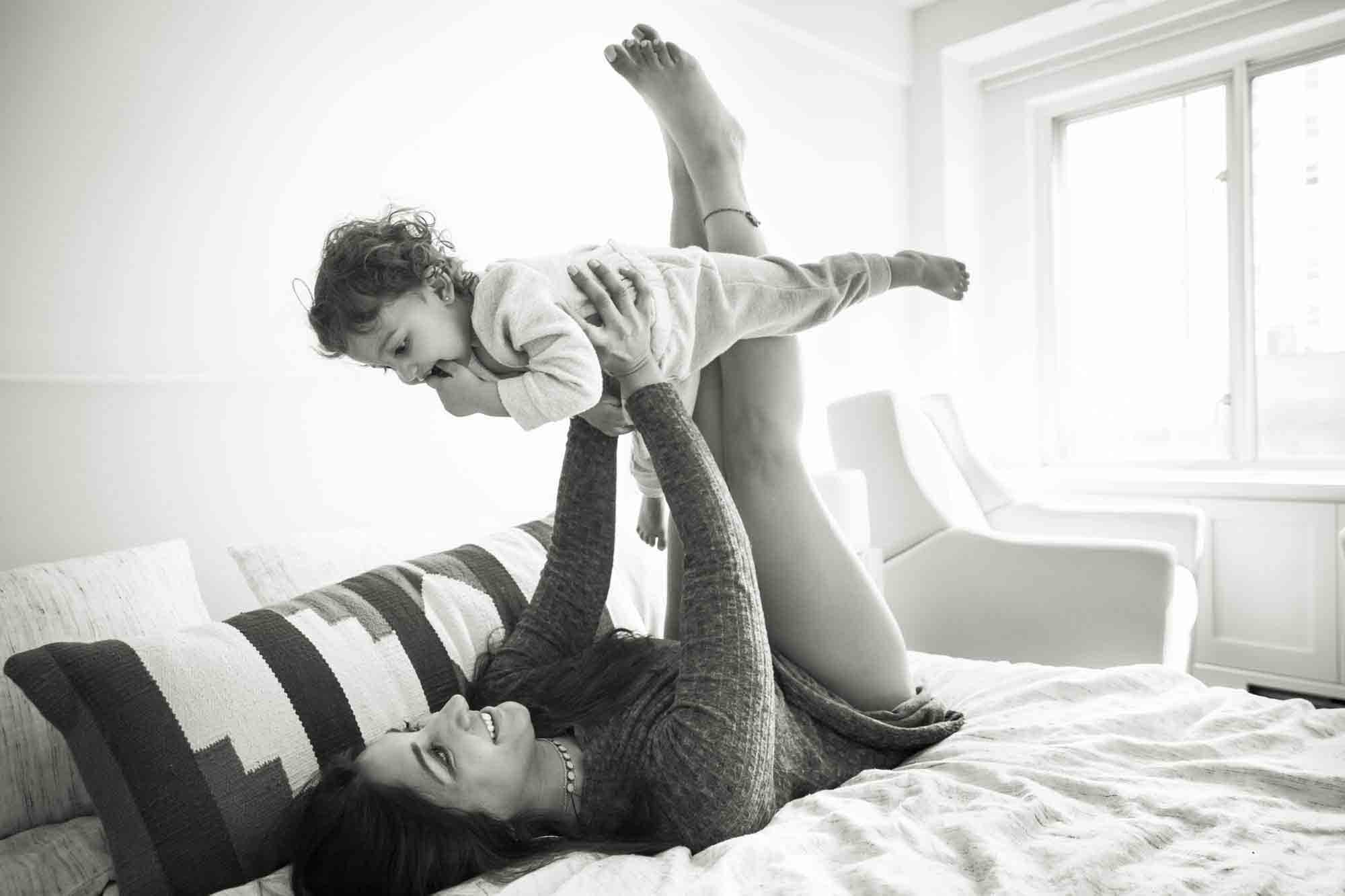 Mother playing with toddler in the air while lying on bed during a NYC family portrait session