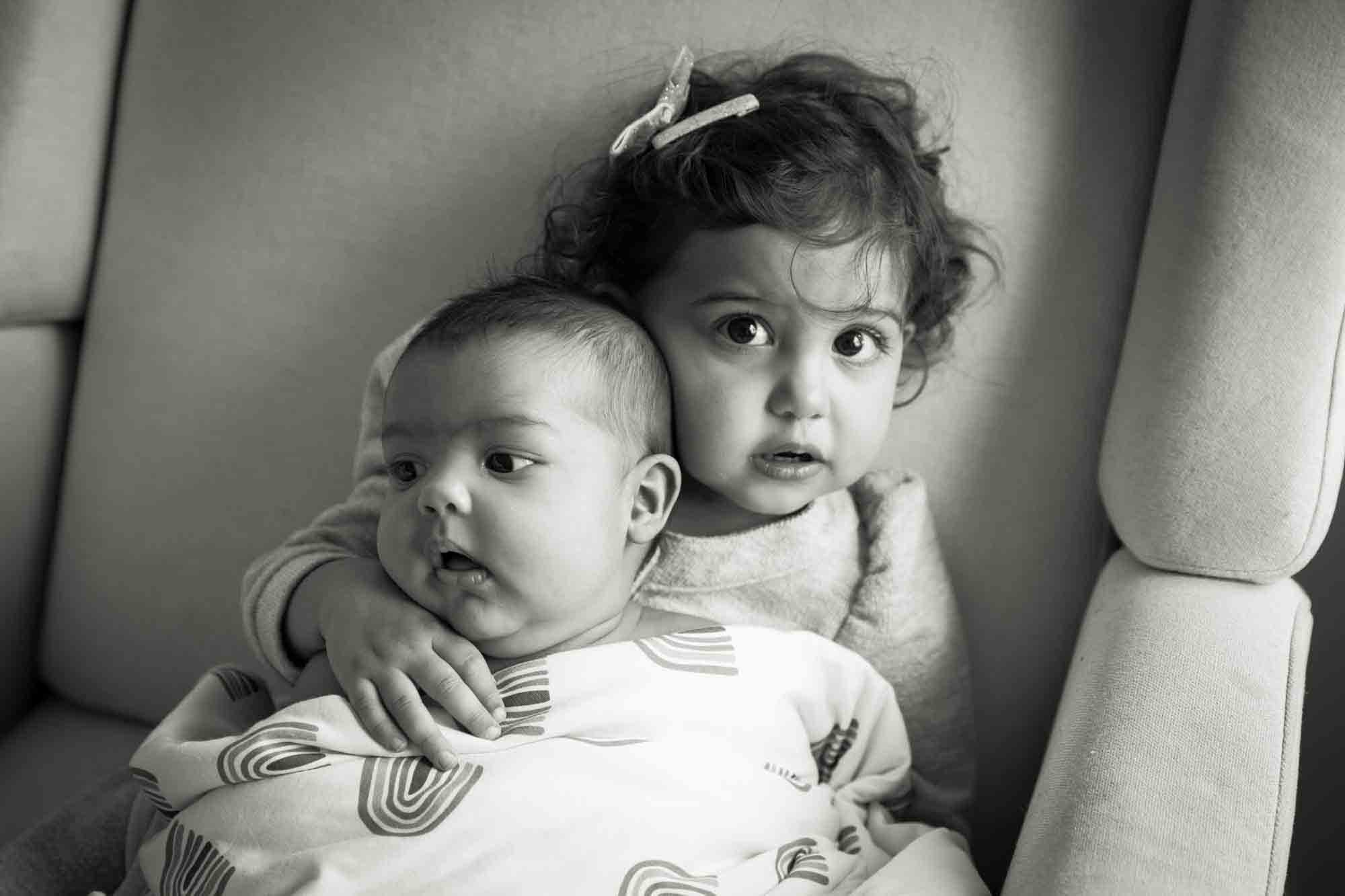 Black and white photo of toddler holding baby in a chair 