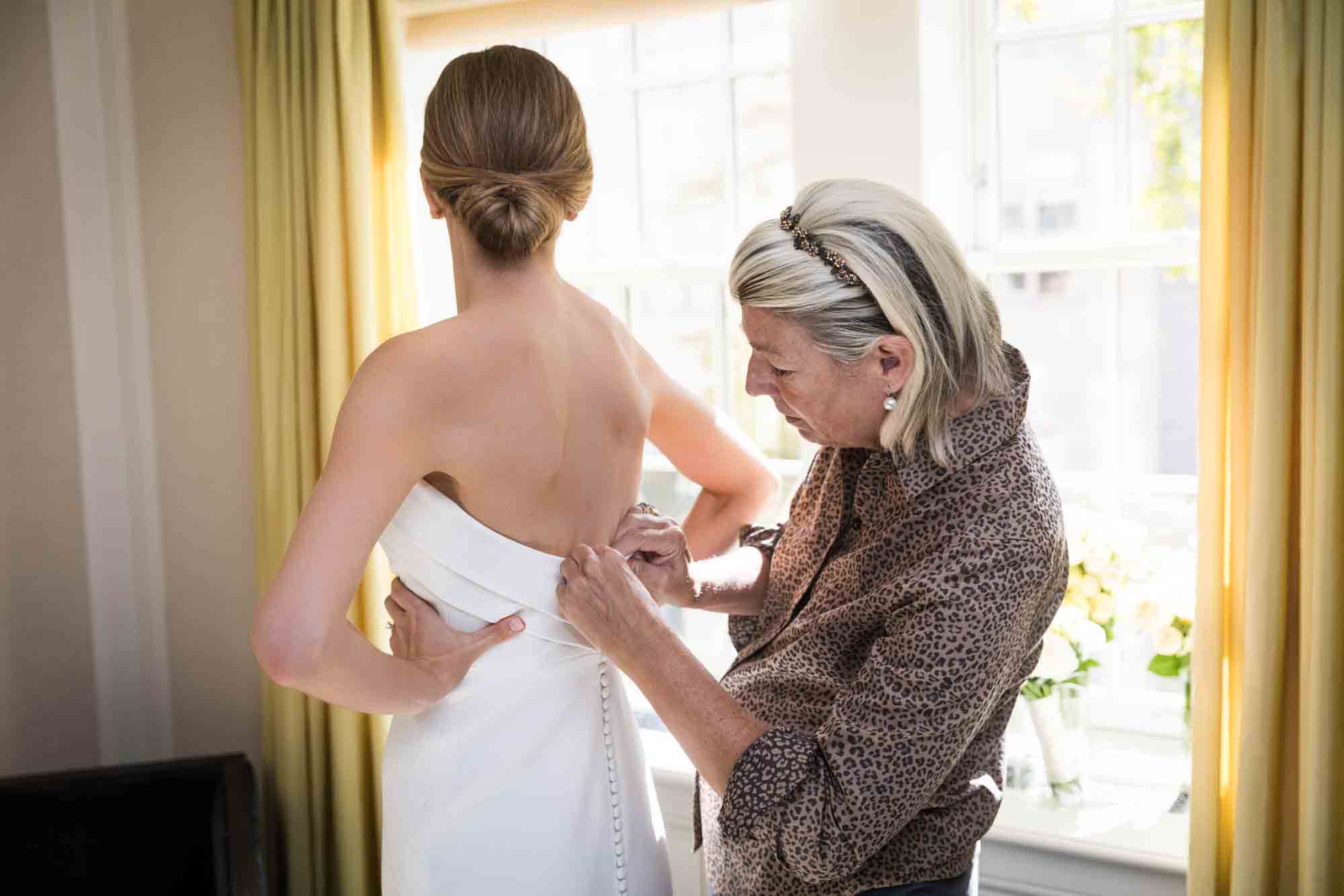 Mother zipping bride's wedding dress in front of window in hotel room