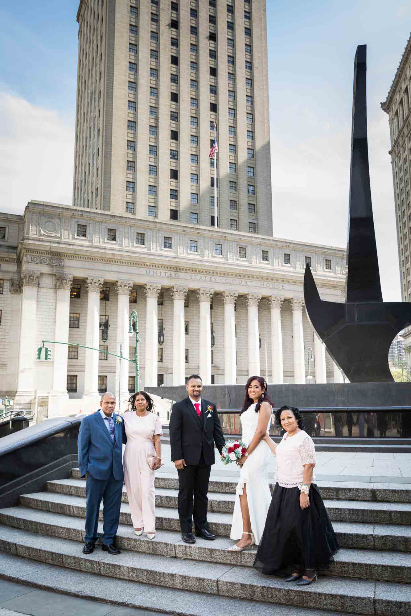 Bride and groom and family on stone steps in front of building and sword statue  at a NYC City Hall wedding