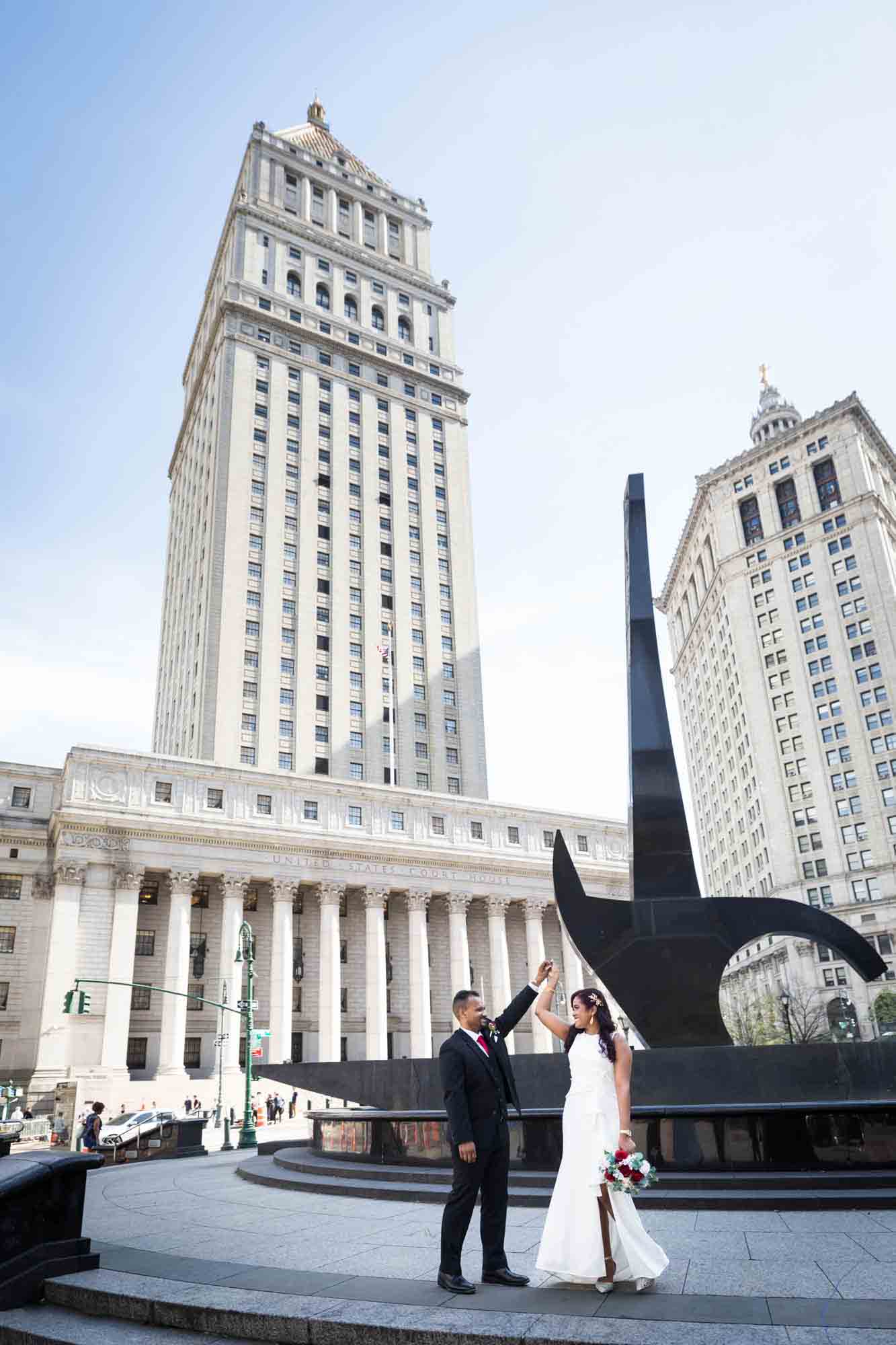 Bride and groom dancing on steps in front of sword statue and large building  at a NYC City Hall wedding